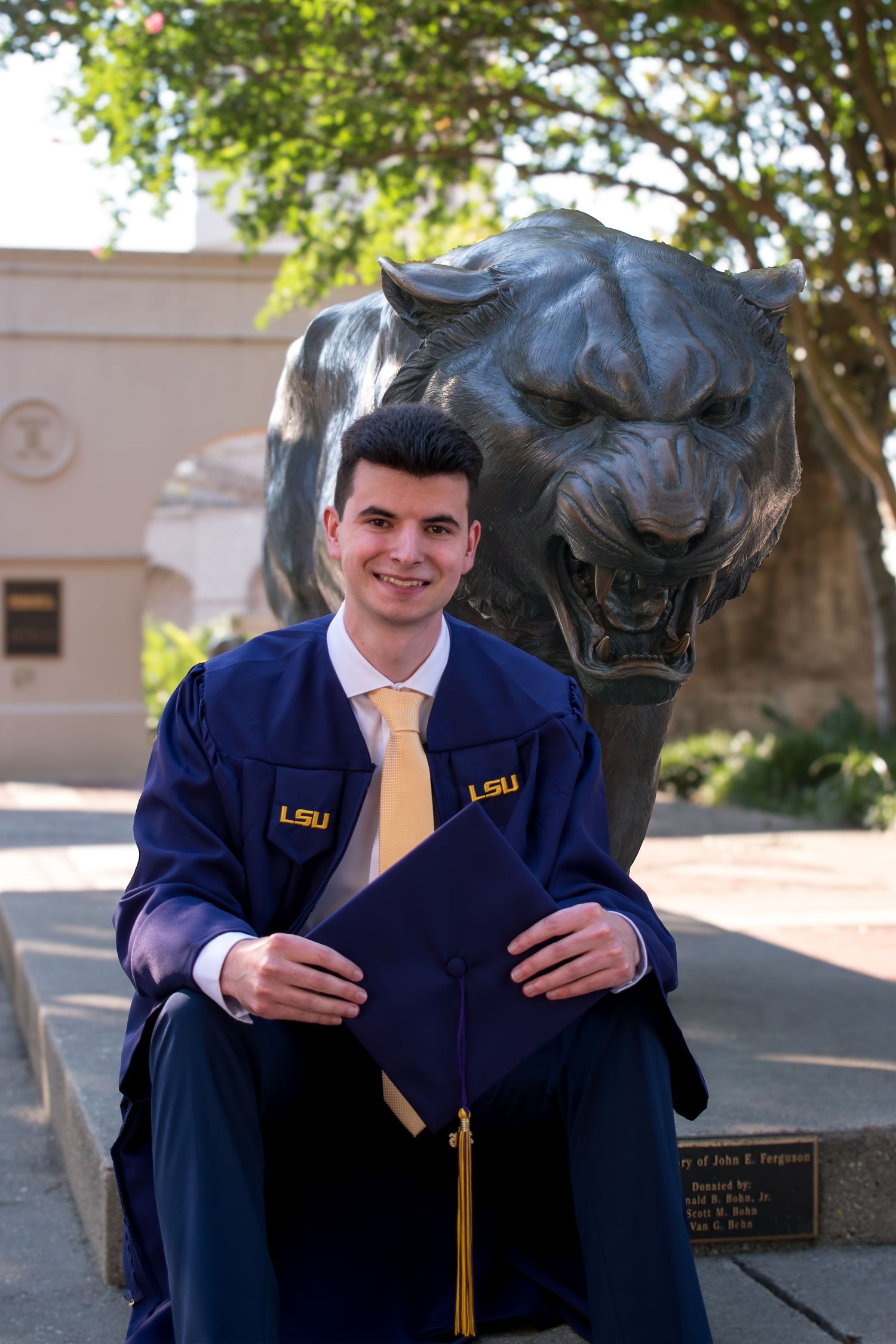 LSU graduate smiling in blue cap and gown, seated in front of the iconic Mike the Tiger bronze statue.