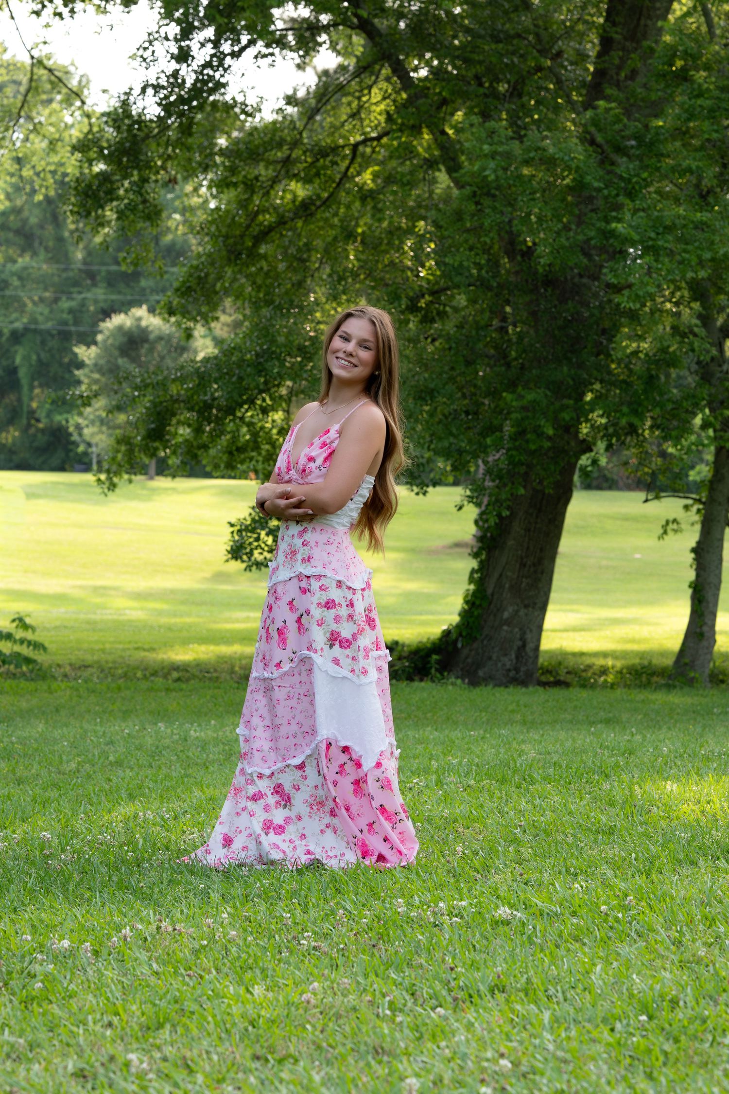 Young woman in pink floral maxi dress smiling outdoors in lush green park setting.