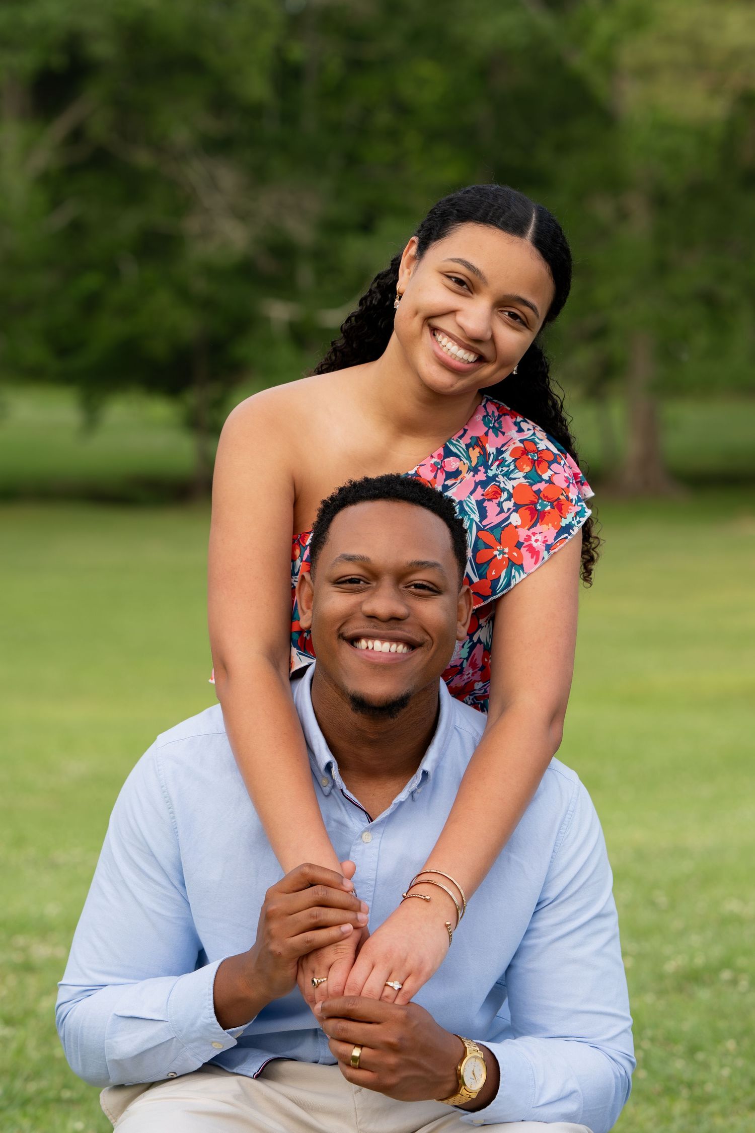 A smiling young Black couple poses outdoors in a park, woman in floral top resting arms on mans shoulders.
