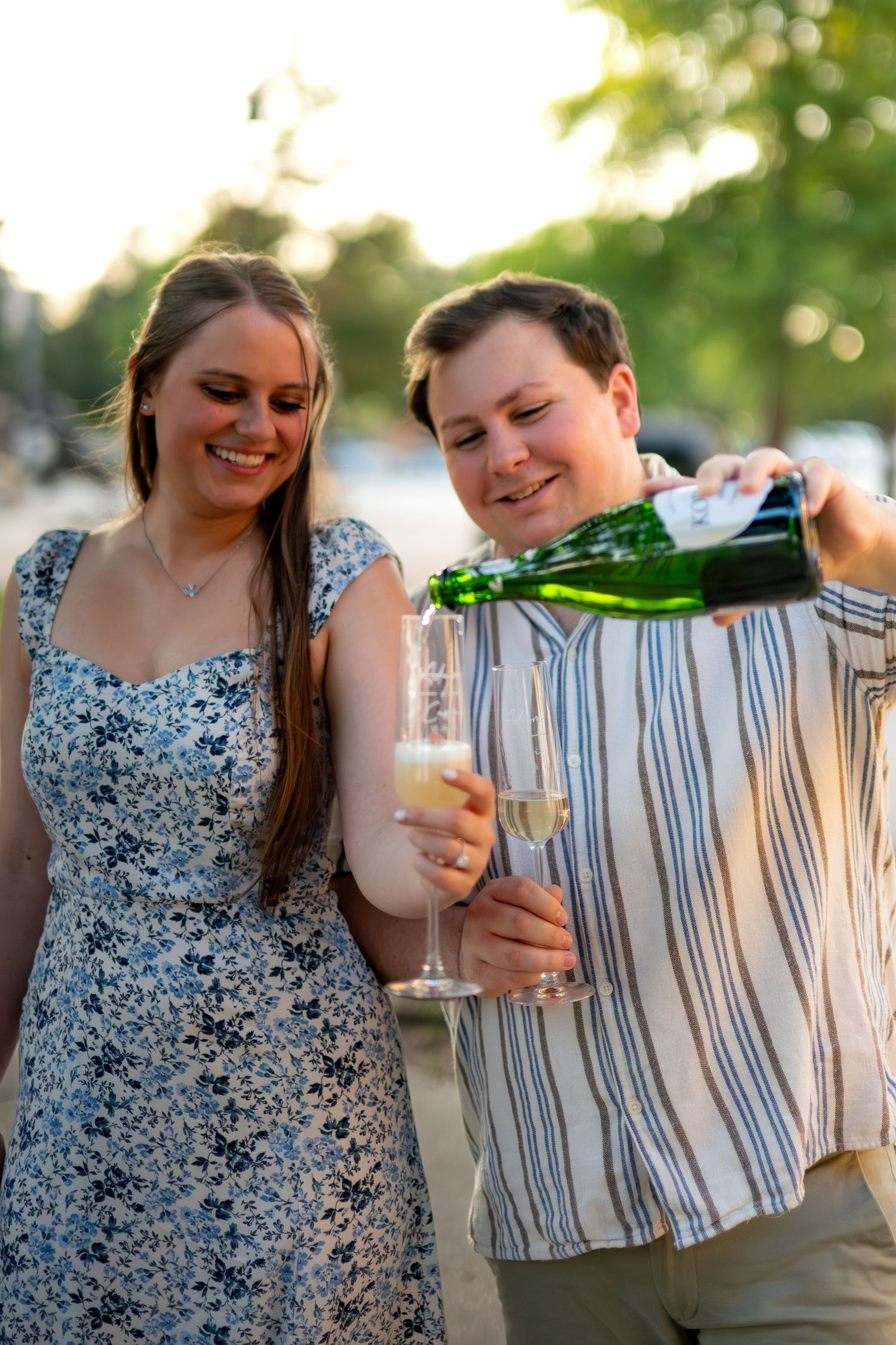 Happy couple celebrating outdoors, man pouring champagne into glasses, woman in floral dress smiling at golden hour.