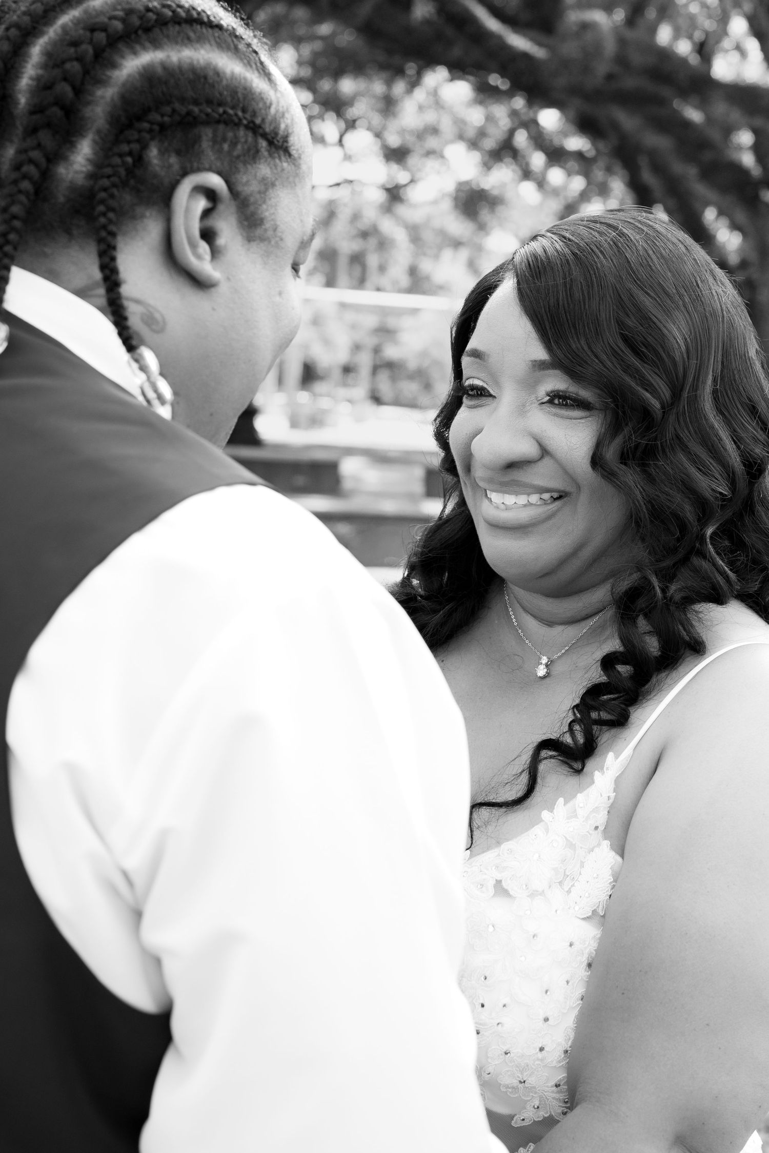 Black and white wedding photo of a smiling bride in lace dress gazing at her partner during an outdoor ceremony.