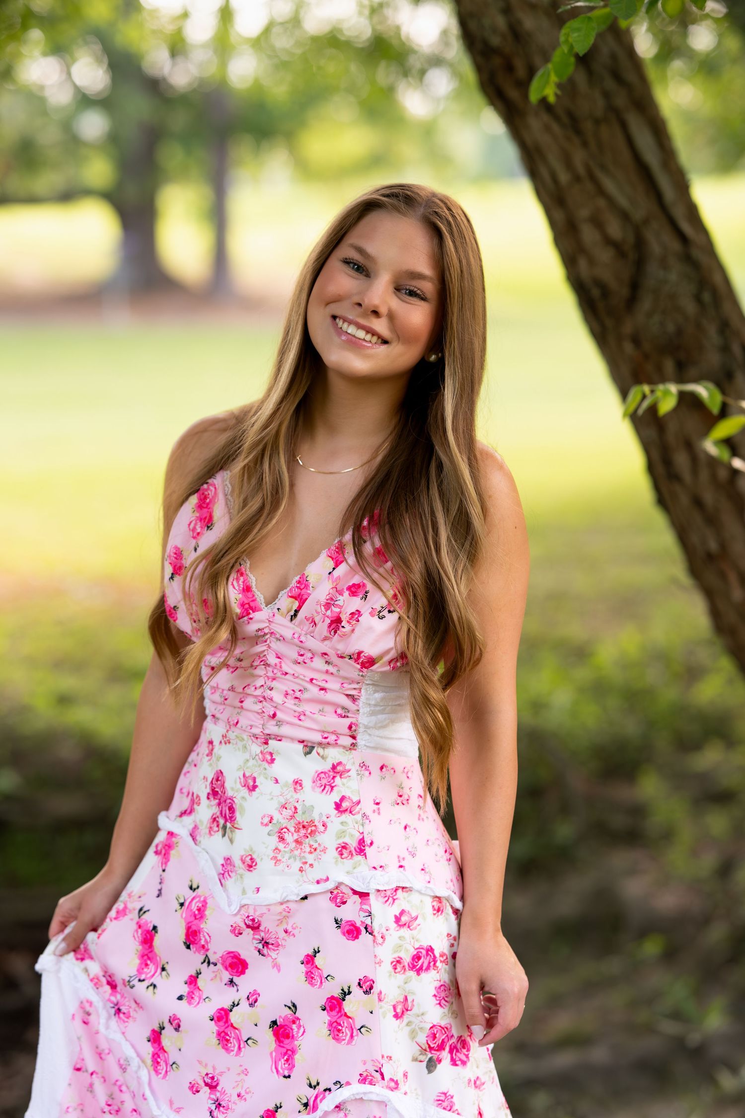 Smiling teenage girl in pink floral dress posing beside a tree in a lush green park.