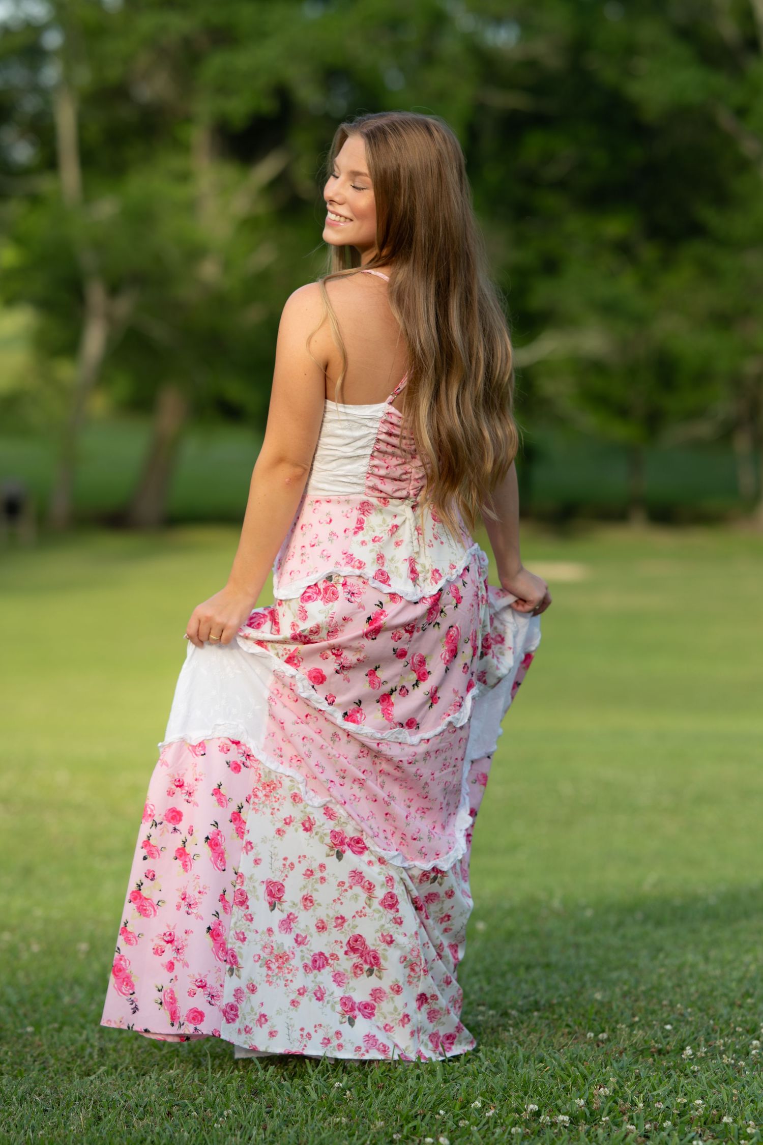 Smiling young woman in pink floral maxi dress posing outdoors in lush green park setting.