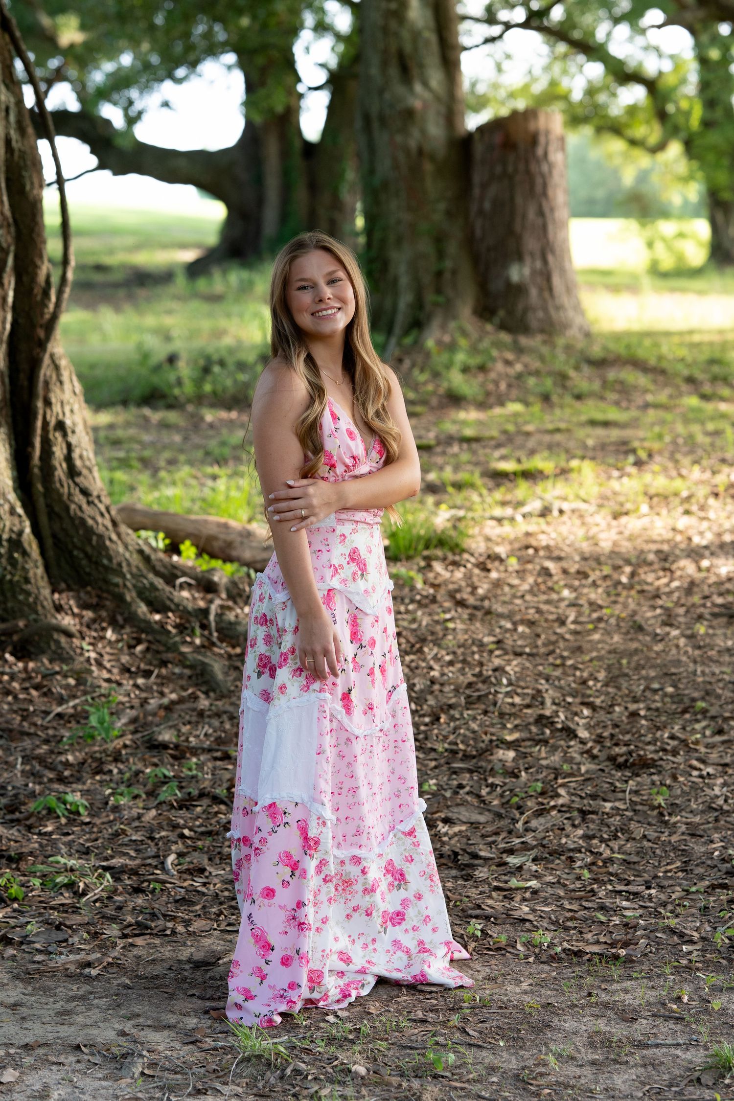 Smiling young woman in pink floral maxi dress stands among large oak trees outdoors.