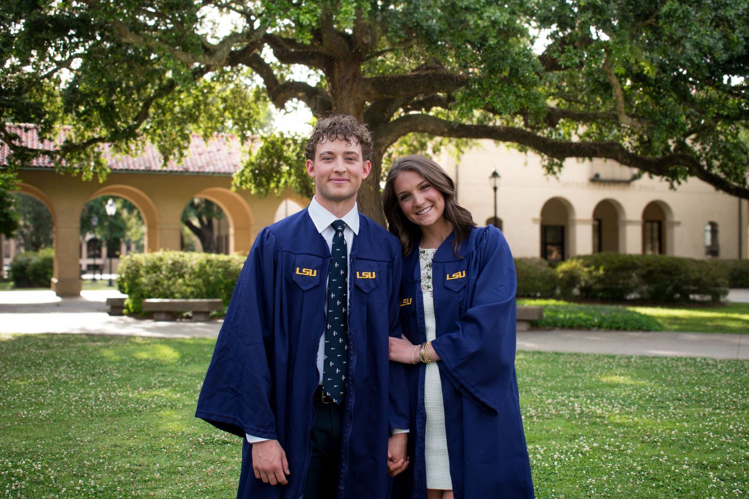 Two smiling graduates in blue LSU caps and gowns pose outdoors on a sunny campus lawn.