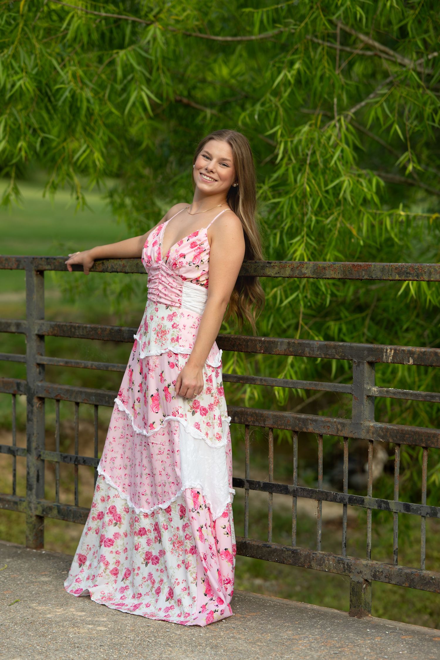 Smiling young woman in pink floral maxi dress posing by a rustic fence with green trees.