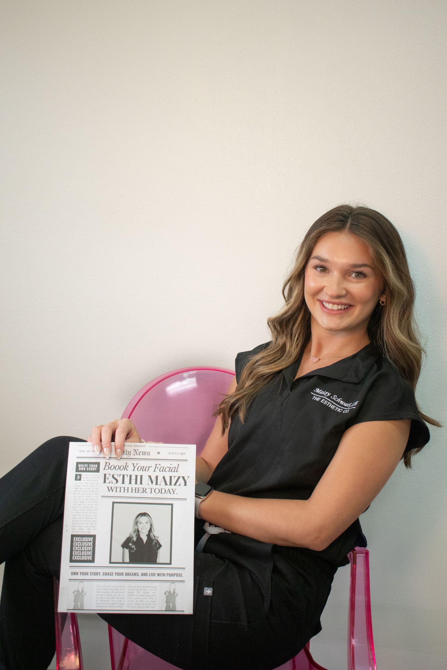 A brunette esthetician in black scrubs sits holding a newspaper-style promotional flyer in a pink chair.