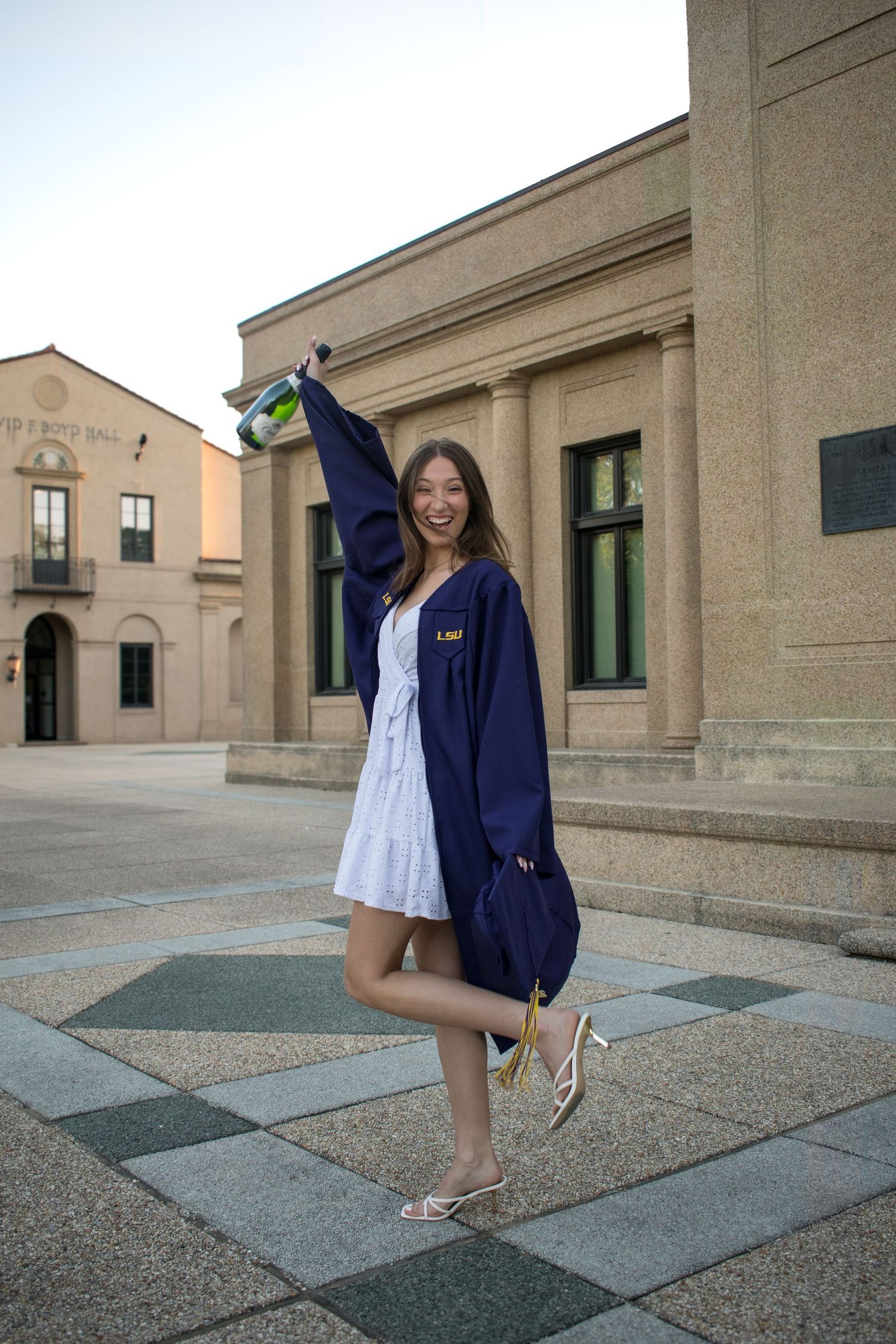 Excited female graduate in navy LSU gown celebrates outdoors raising cap near campus building.