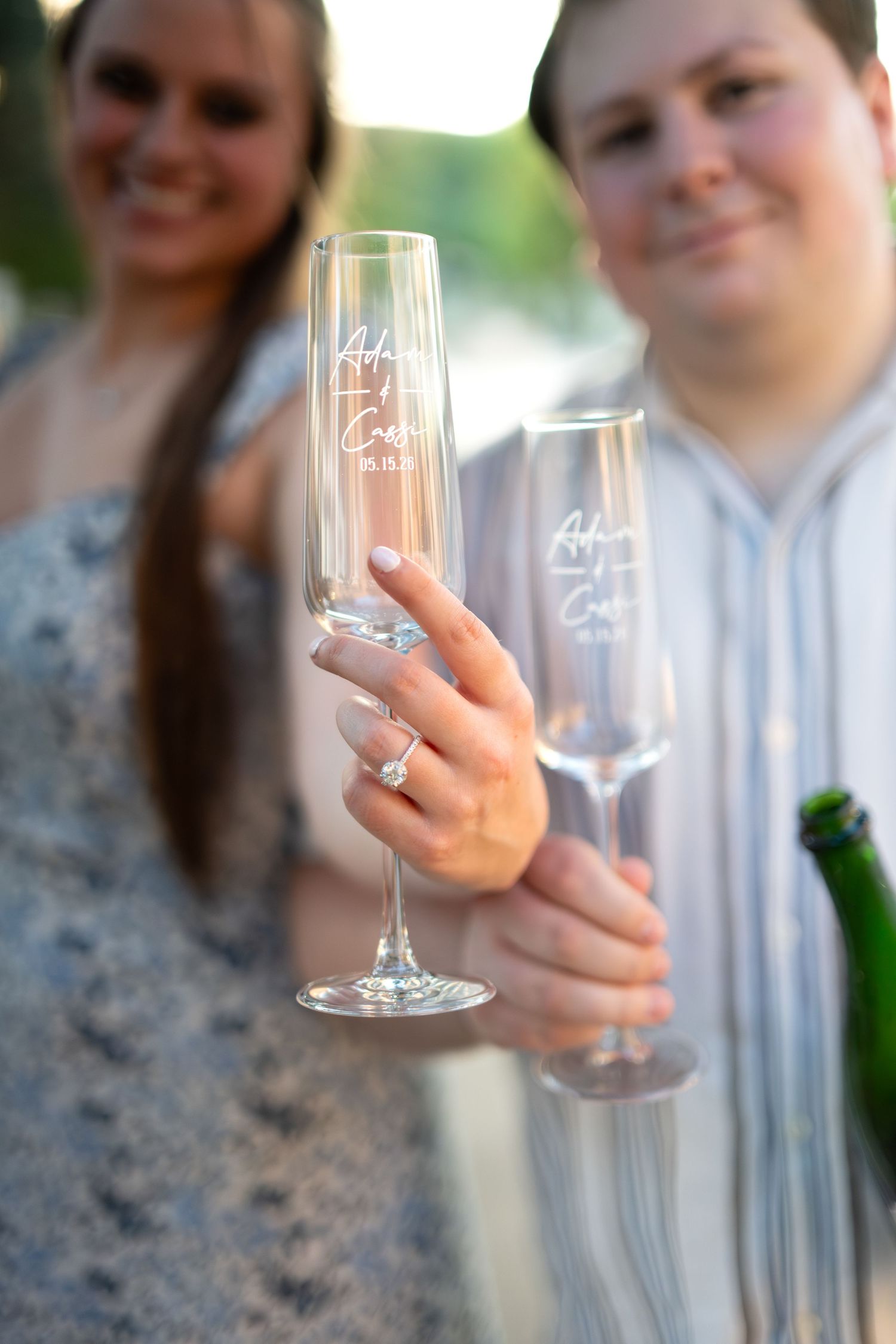 Engaged couple holding personalized champagne flutes with engagement ring visible outdoors.