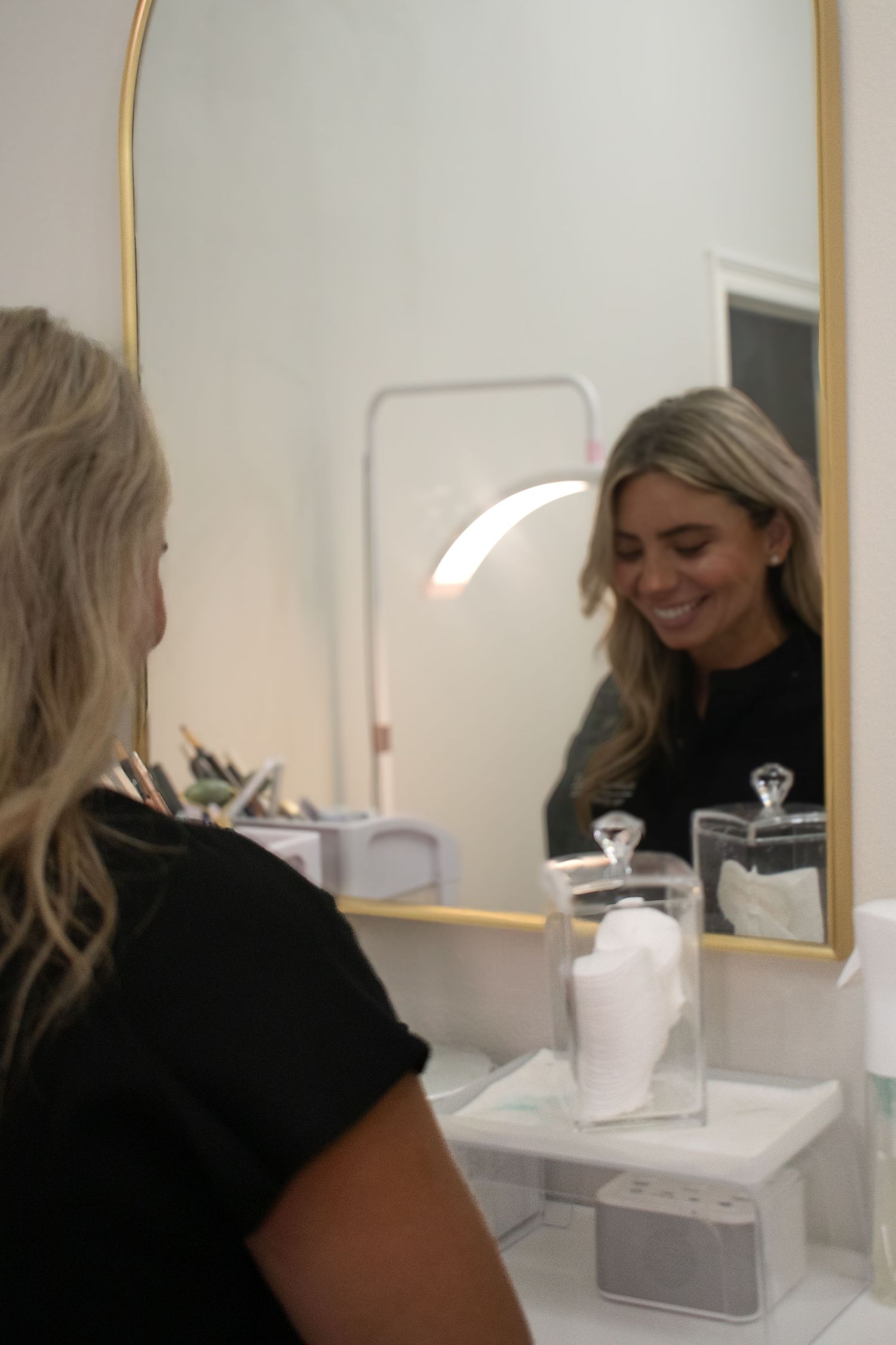 A blonde esthetician in a black top smiles at her reflection in a gold-framed bathroom mirror.