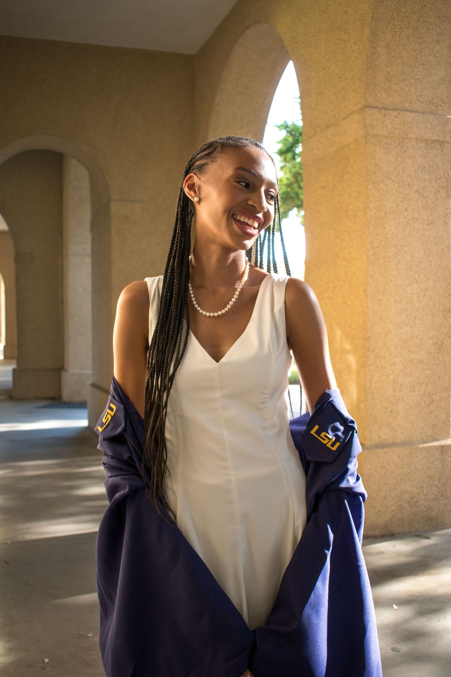 Smiling Black woman with braids in white dress holding LSU graduation gown in sunlit archway.