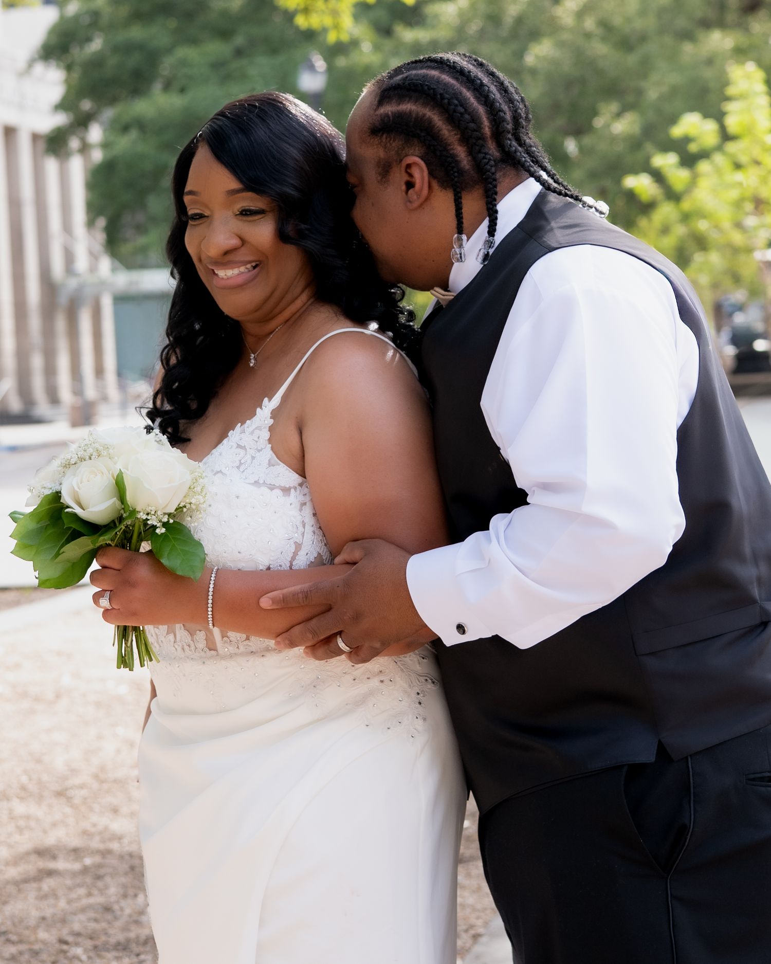 A smiling bride in a white lace gown holds a bouquet while her groom in a black vest kisses her cheek outdoors.