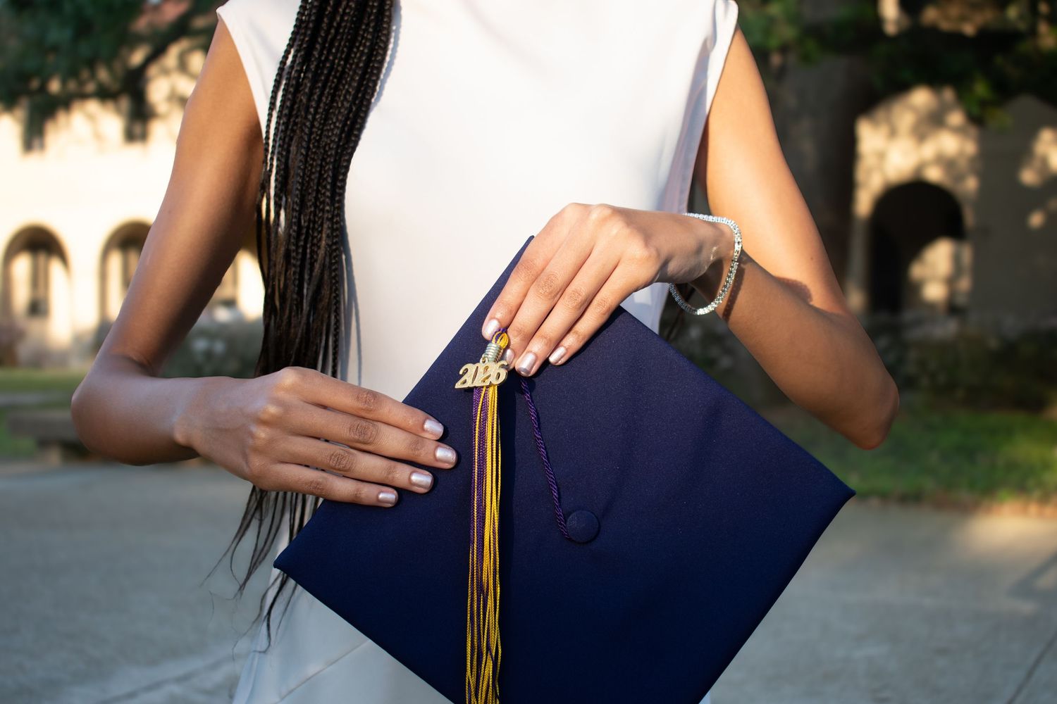 A Black woman in a white dress holds a navy blue graduation cap with gold tassel outdoors.