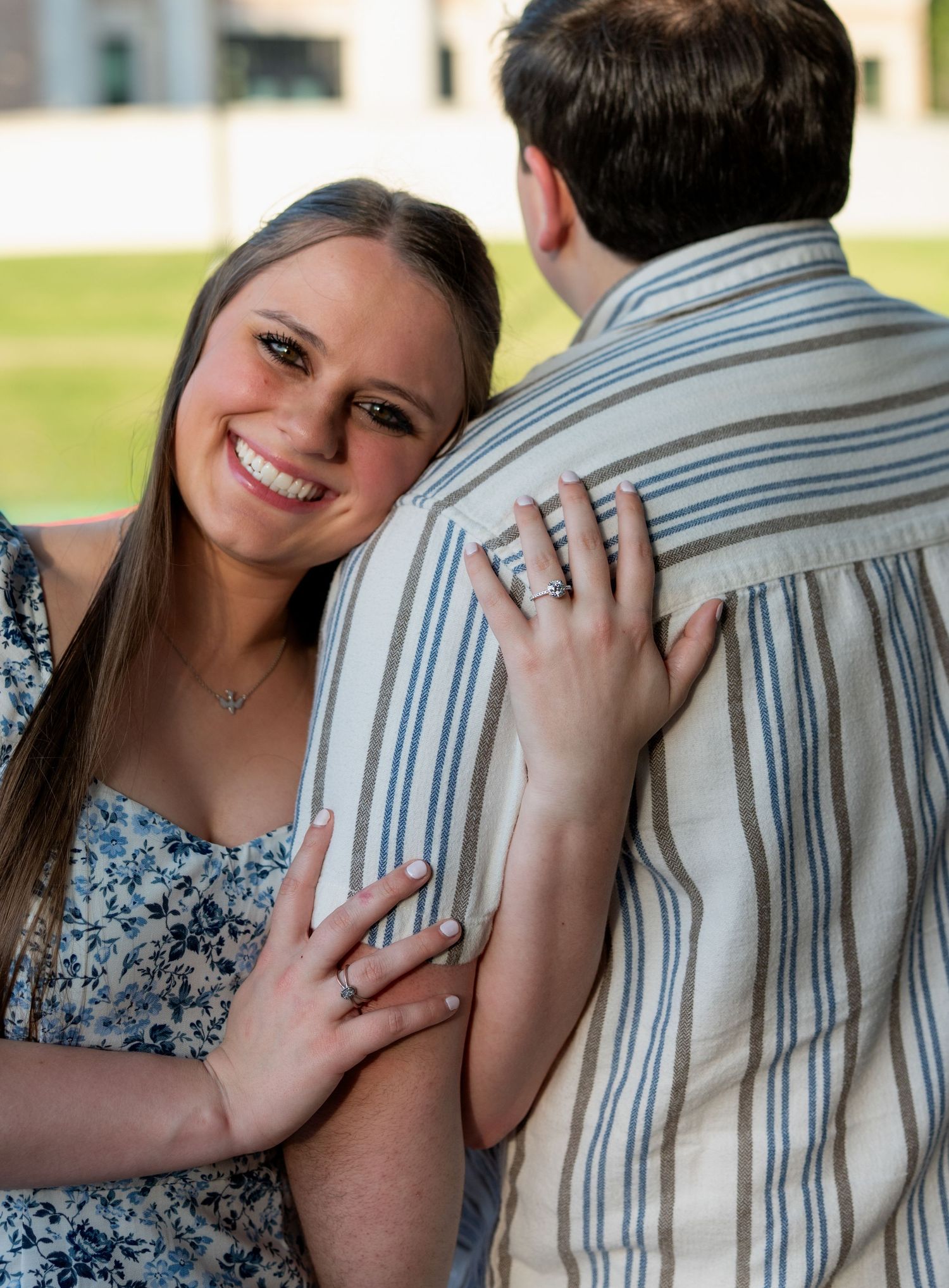 Smiling woman in floral dress hugs man from behind, showing engagement ring, outdoors in soft natural light.