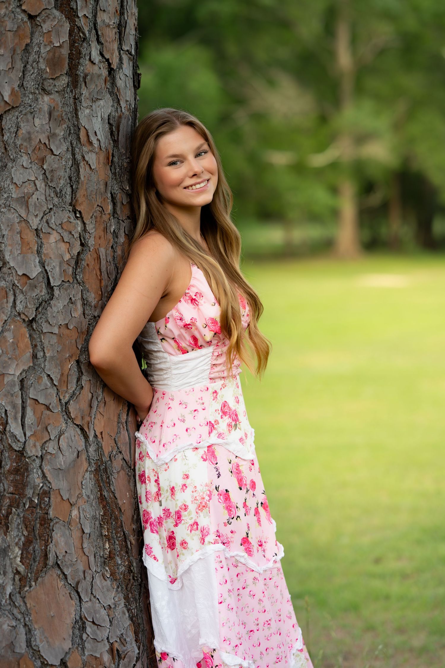 Smiling young woman in pink floral maxi dress leaning against a pine tree in a park.