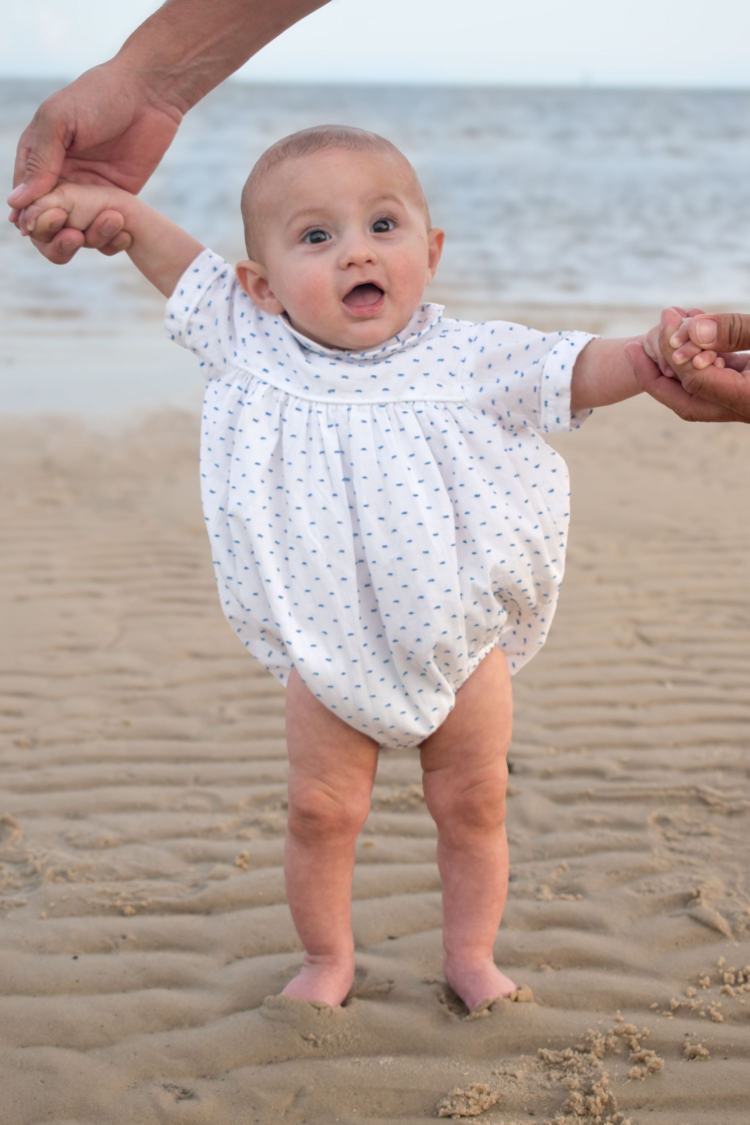 Adorable baby in white polka dot outfit takes first steps on sandy beach held by hands.