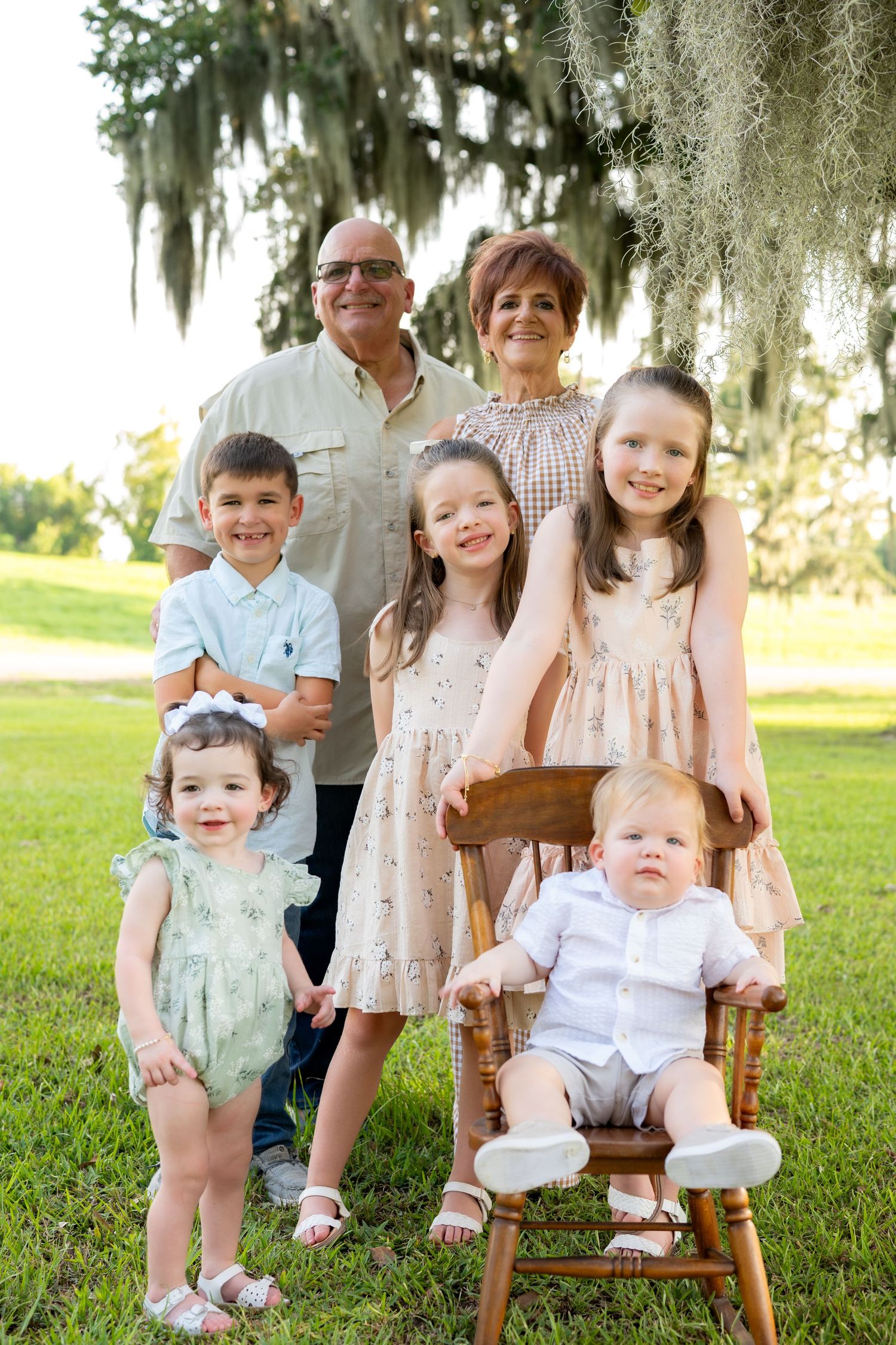 Grandparents pose outdoors with five young grandchildren, one seated on a wooden rocking chair, beneath a mossy oak tree on green grass.