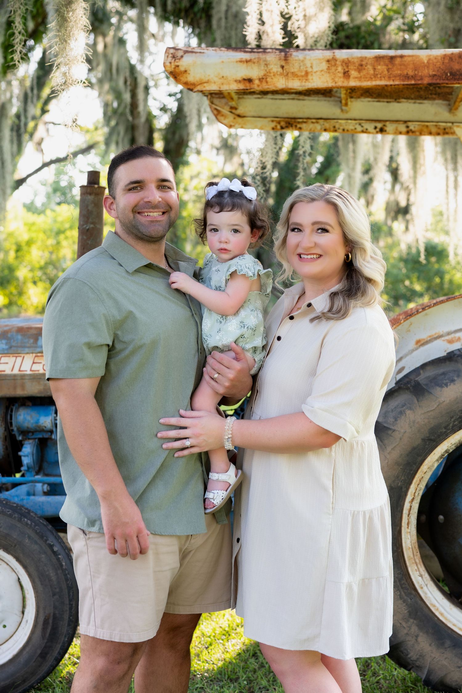 A smiling family of three poses in front of a vintage tractor under moss-draped trees outdoors.