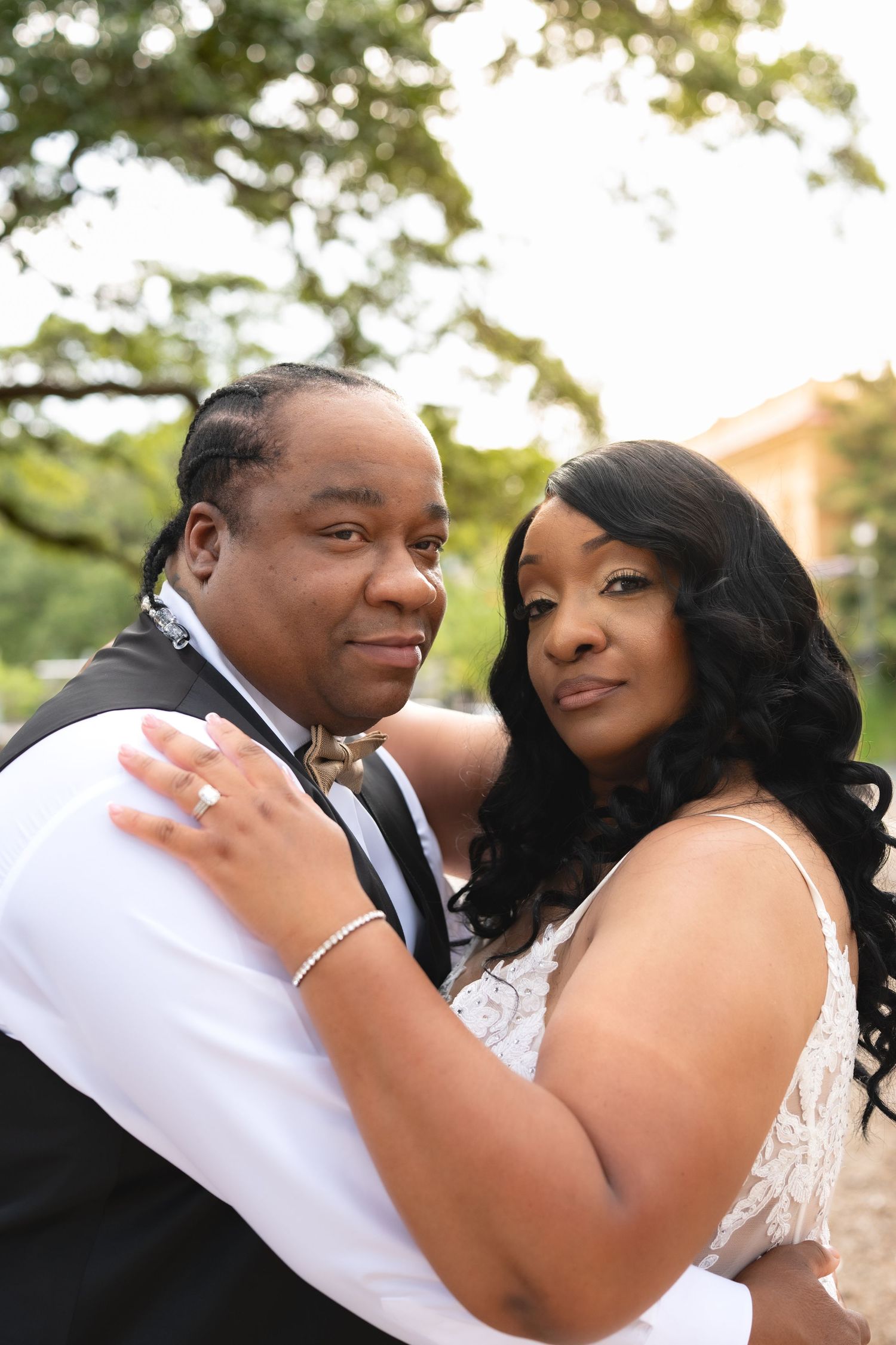 A couple in wedding attire embraces outdoors, the bride in lace and groom in a suit, posing for a romantic portrait.