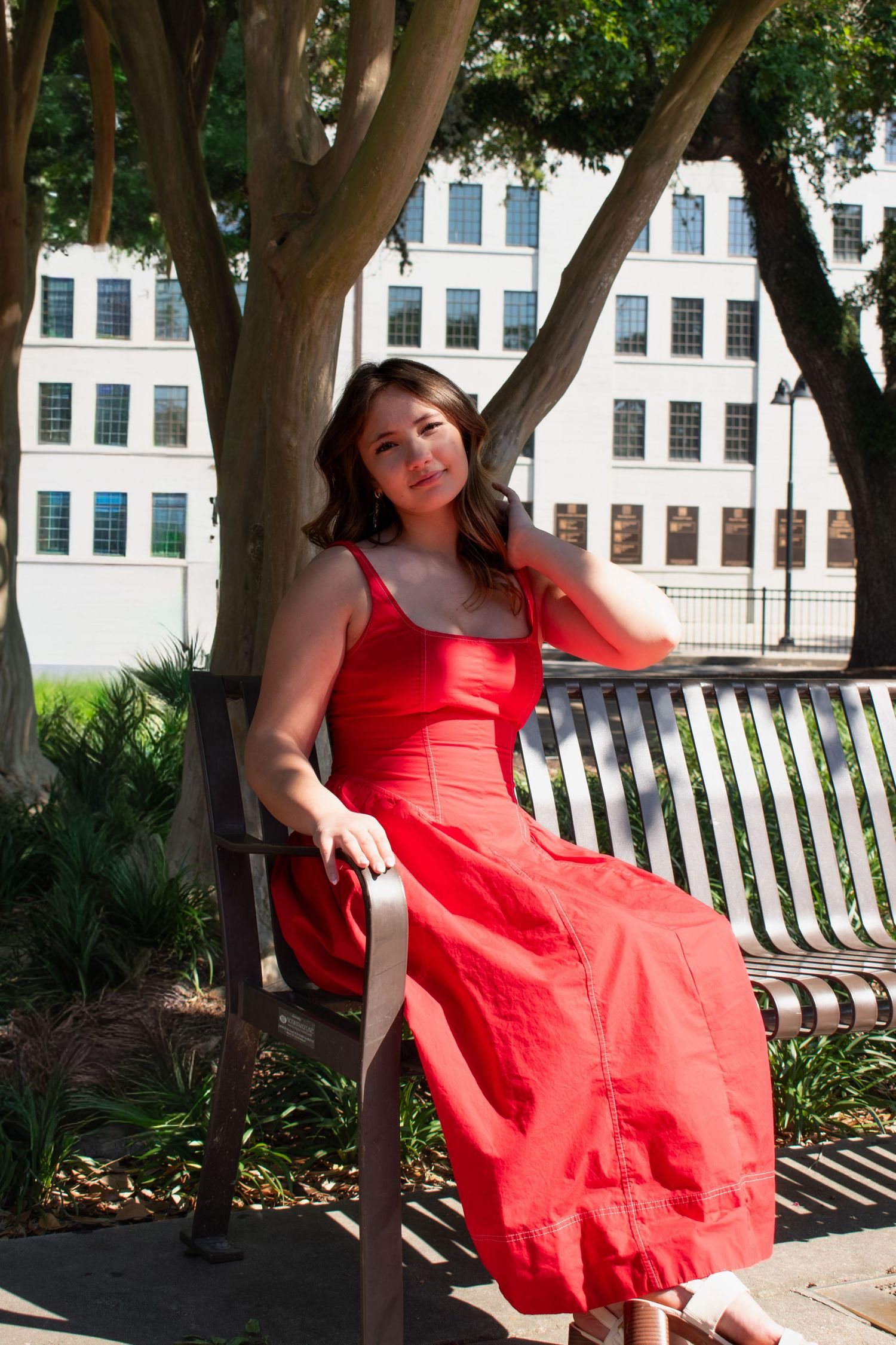 Woman in red maxi dress sitting gracefully on a park bench with trees and white building backdrop.