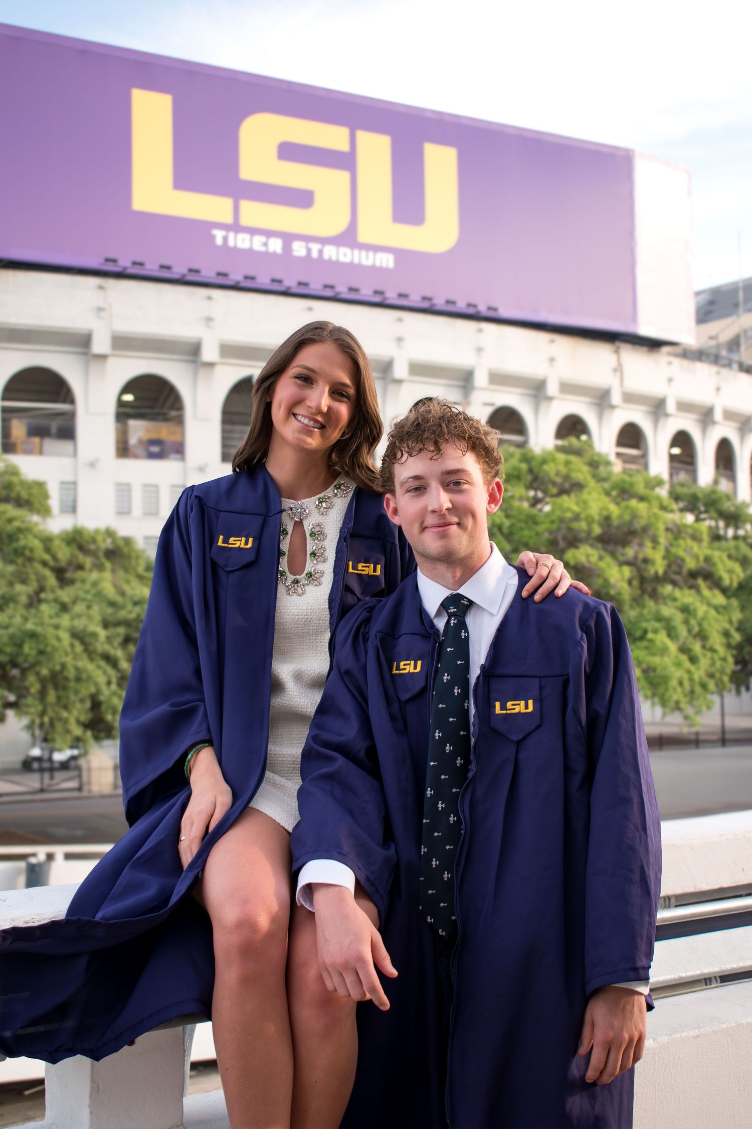 Two LSU graduates in navy blue graduation gowns pose proudly in front of Tiger Stadium at Louisiana State University.