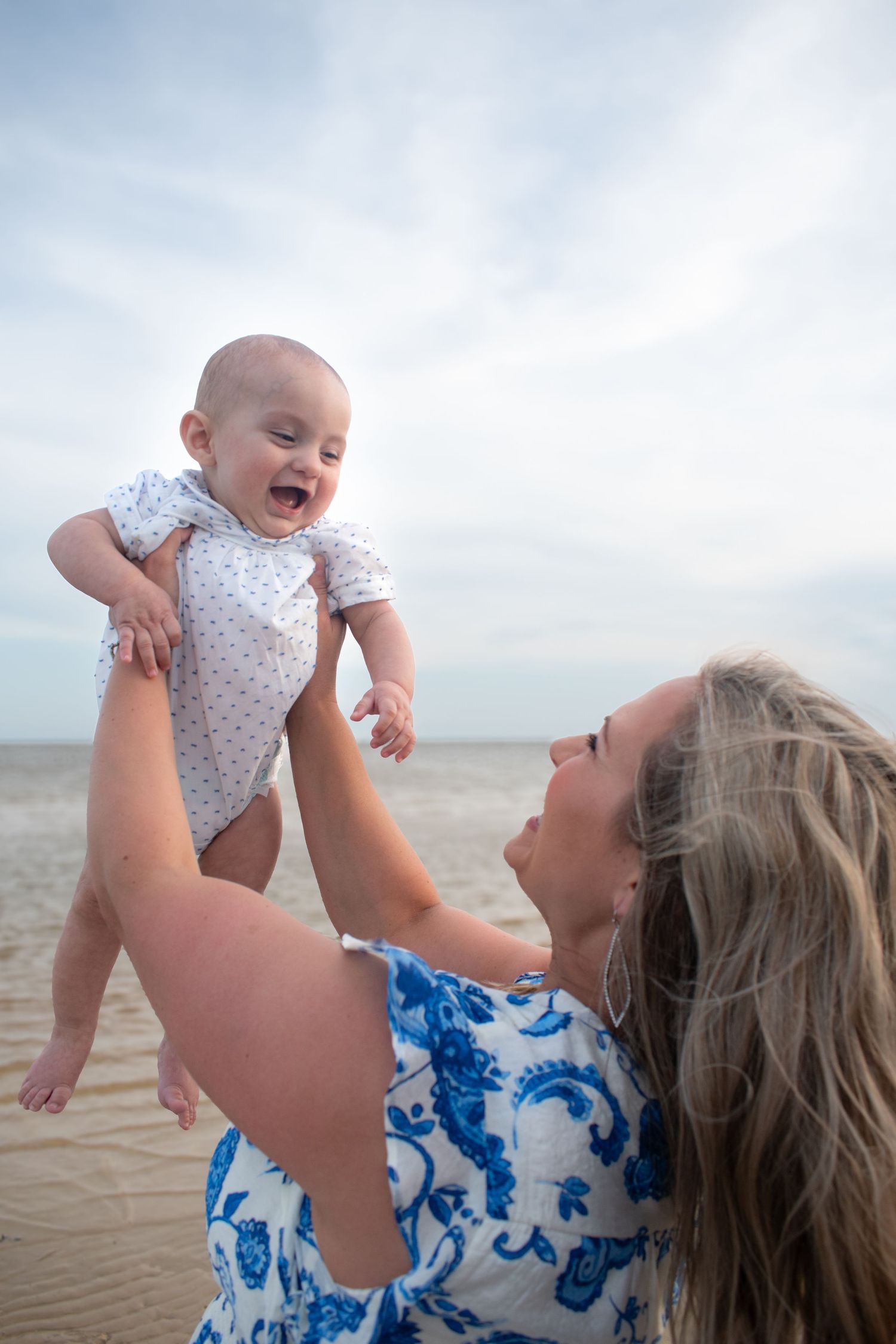 Joyful mother lifts laughing baby high in the air on a breezy beach at sunset.