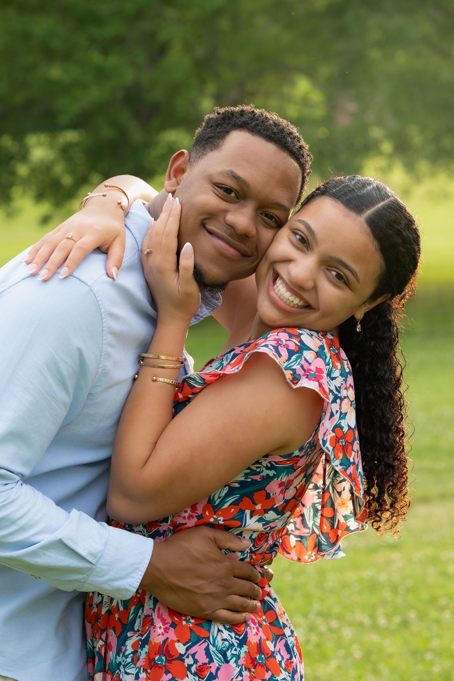 Smiling couple hugging warmly outdoors, woman in floral dress embracing man in light blue shirt.