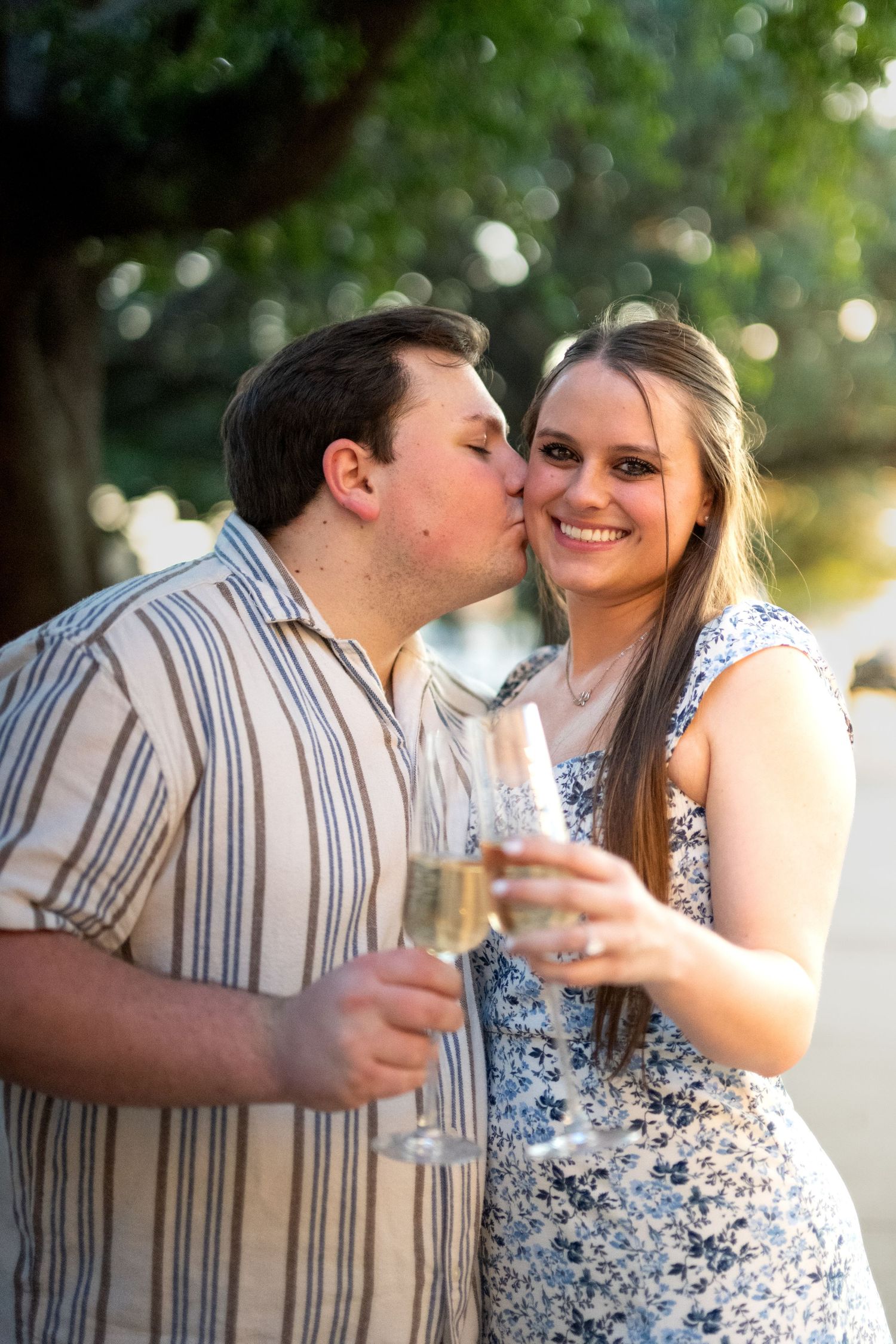 Couple celebrating with champagne glasses as man kisses woman on cheek outdoors near trees.