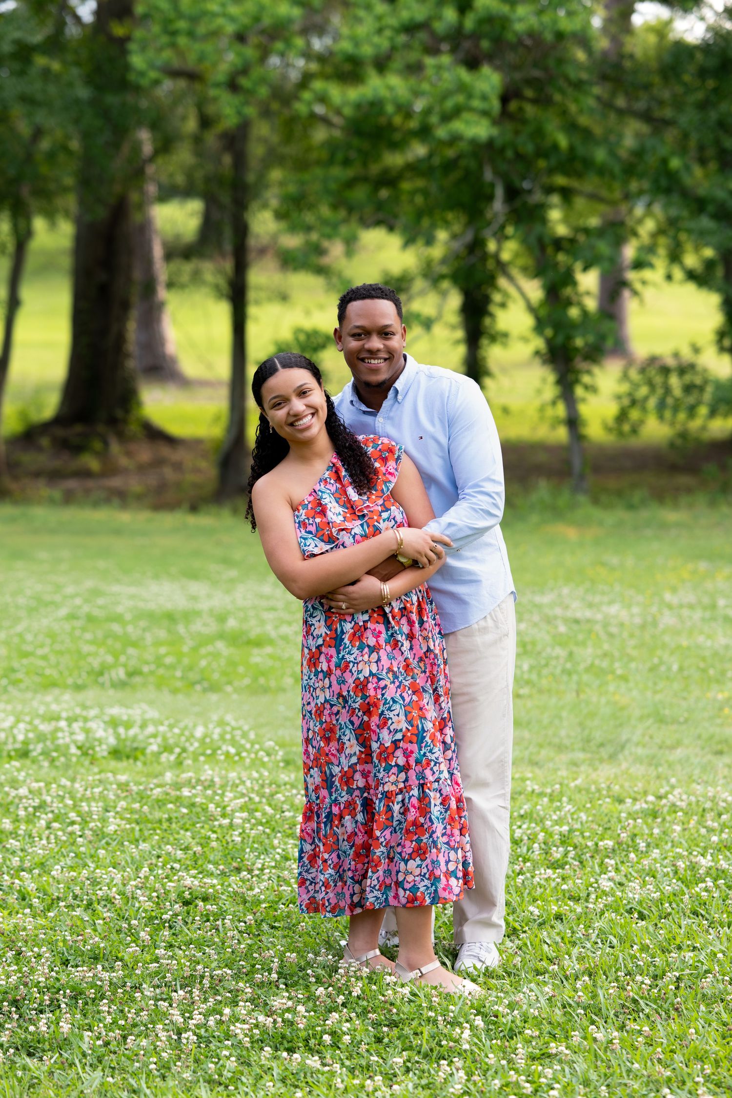 Smiling couple posing together in a flower-dotted meadow, man embracing woman in red floral dress.