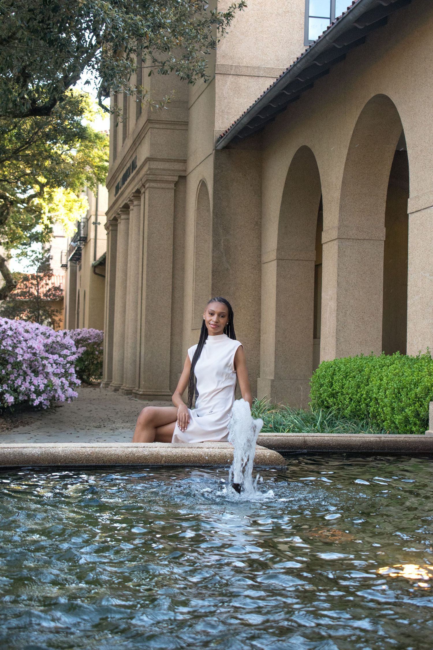 Young woman in white dress sits gracefully beside a decorative fountain with arched building backdrop.