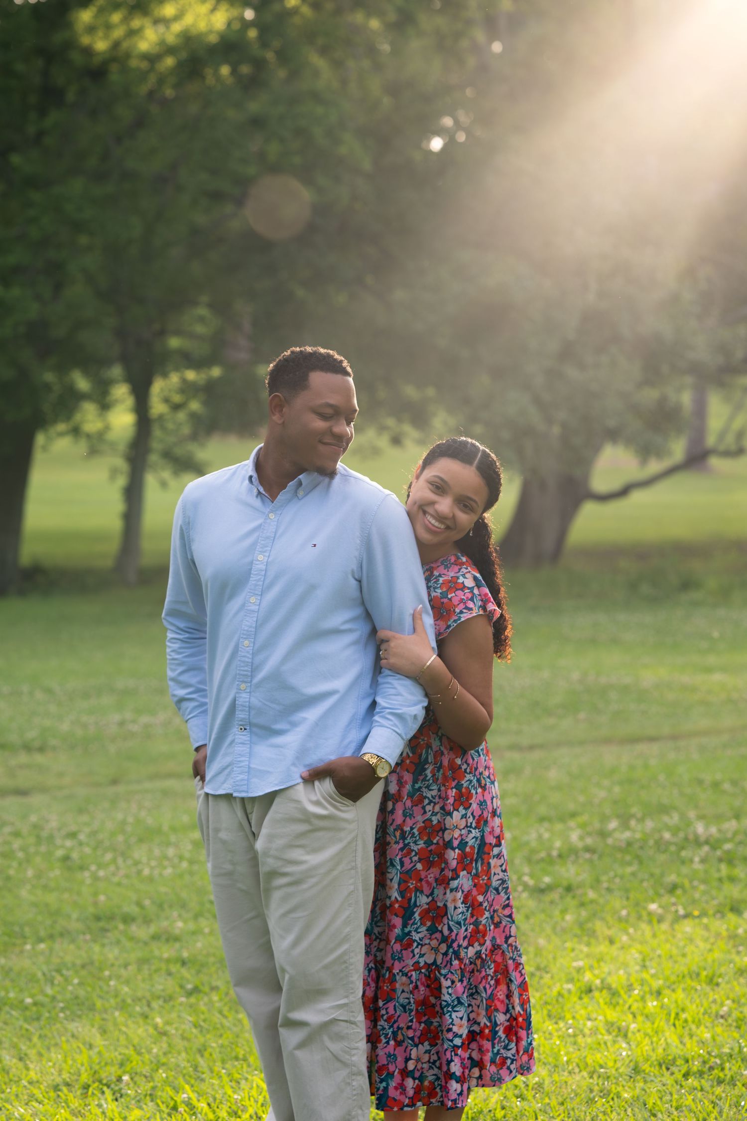 A joyful couple embraces in a sunlit park, woman in floral dress leaning on mans arm in blue shirt.