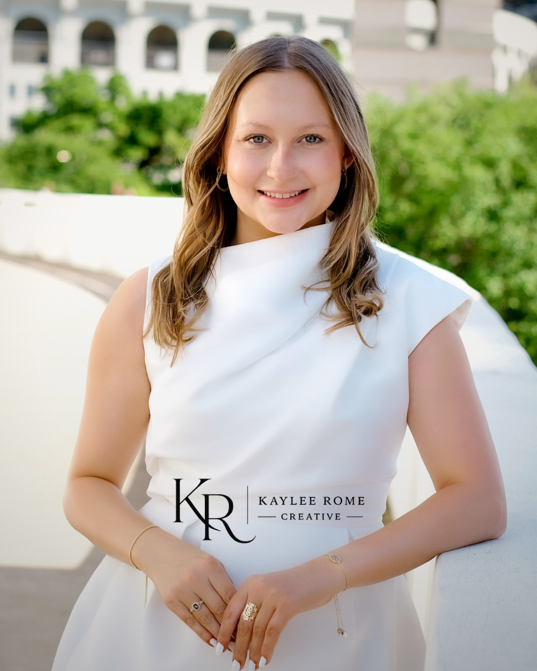 Young woman in white sleeveless dress smiles confidently outdoors in professional portrait photo.