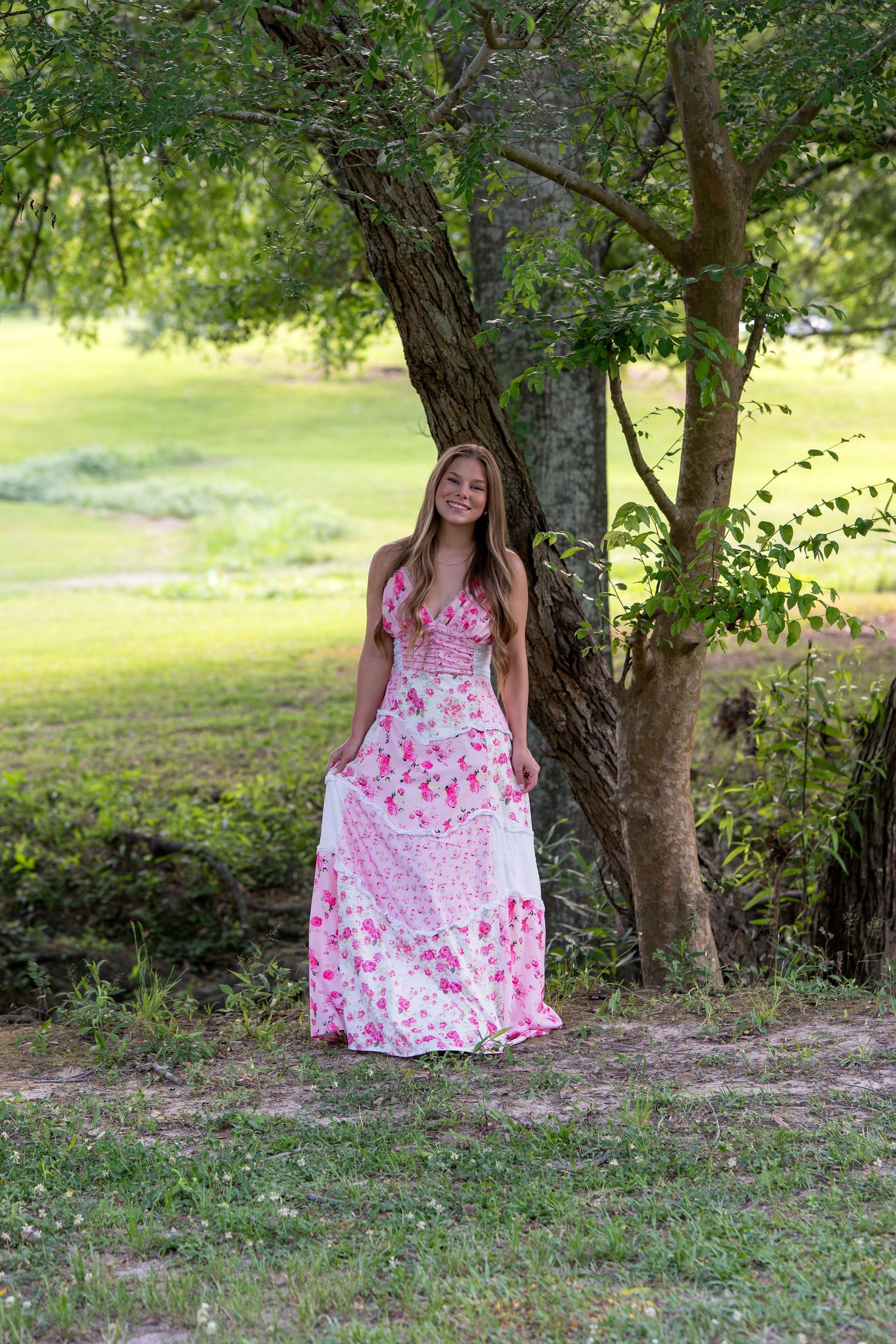 Smiling young woman in pink floral maxi dress leaning against tree in sunlit garden.