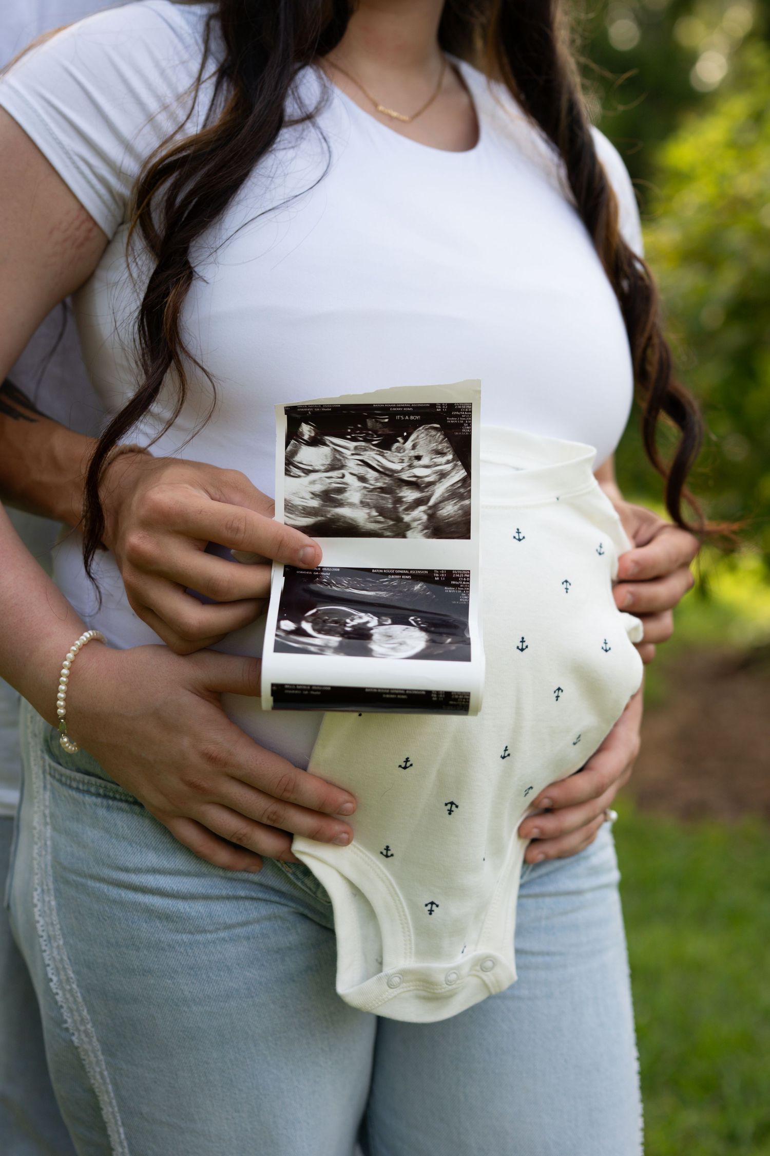 Pregnant woman holding baby ultrasound photos and a white anchor onesie against her baby bump outdoors.
