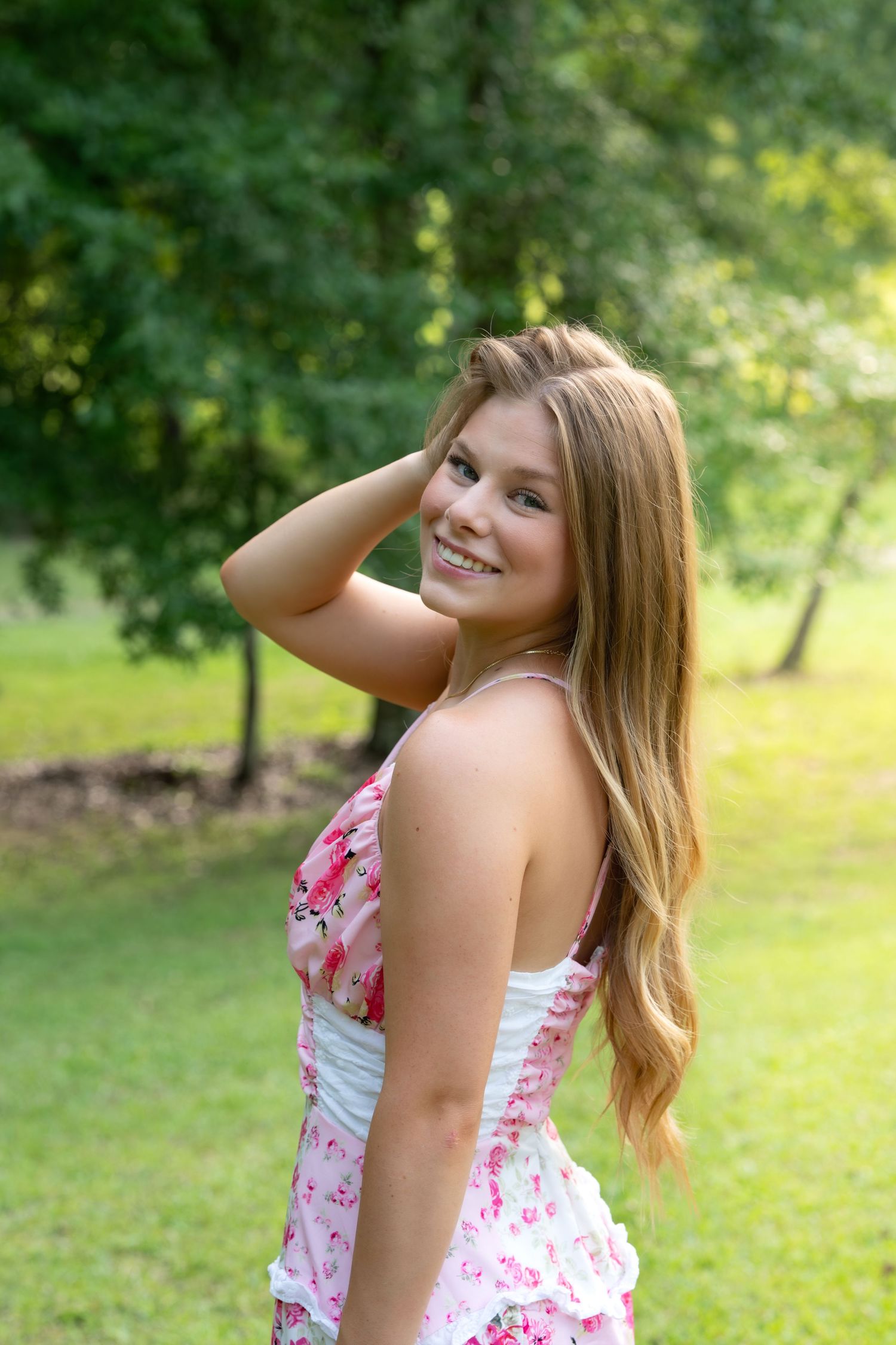 Smiling teenage girl in pink floral dress touching her hair in a sunny green park.