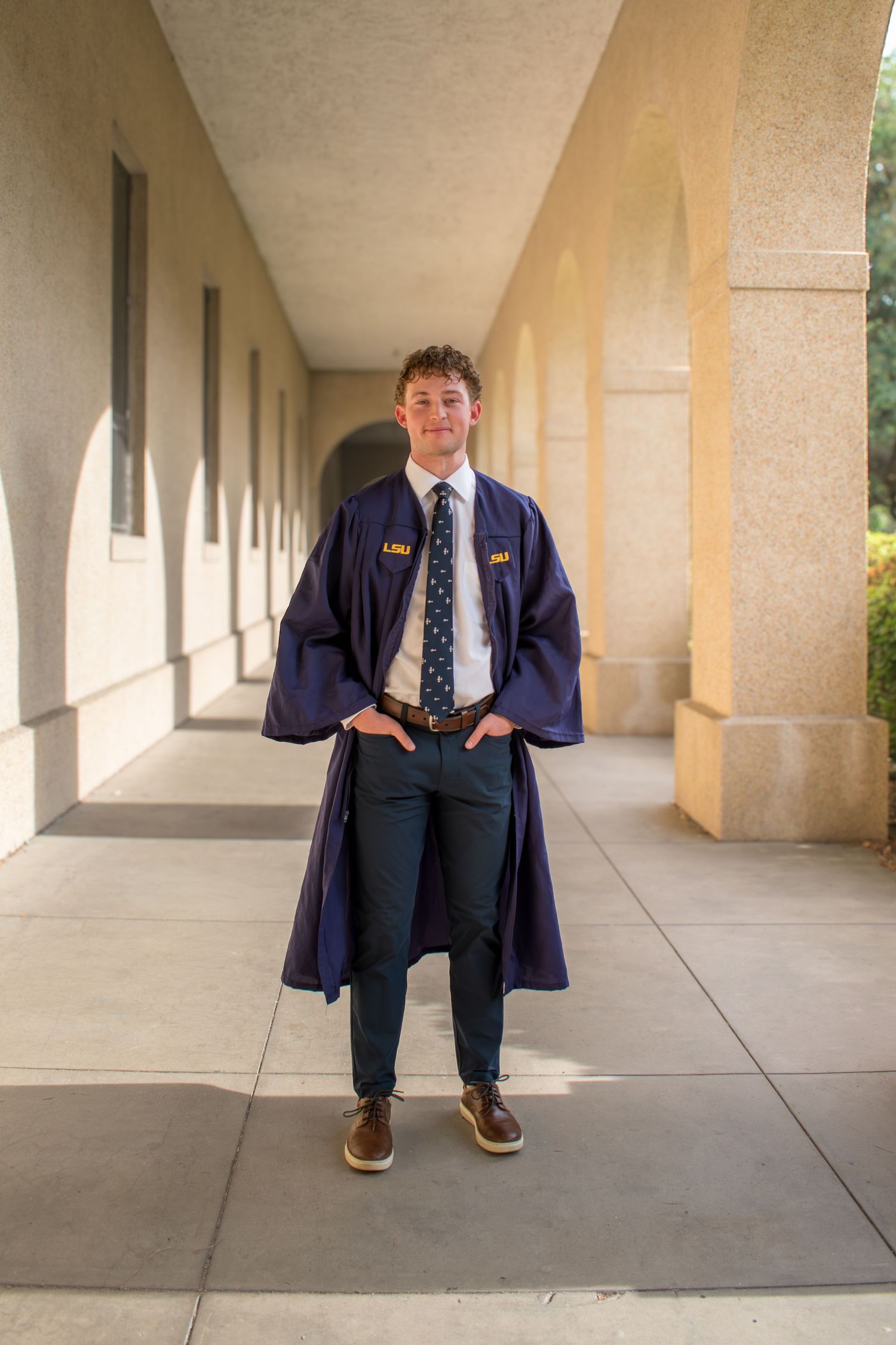 Young man in navy graduation gown and tie poses confidently in a sunlit campus corridor.