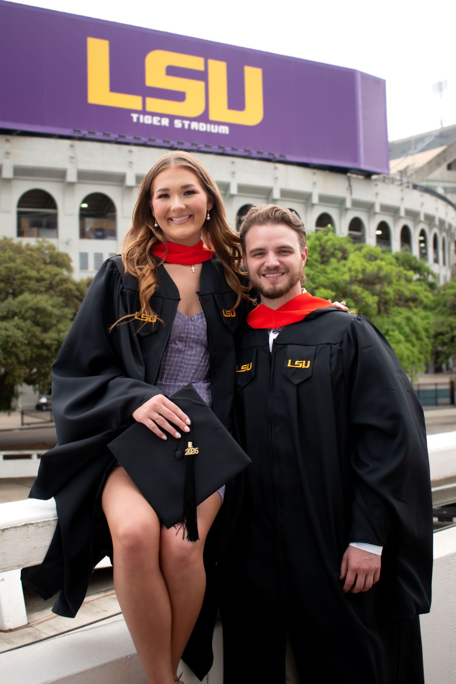 Two LSU graduates in black robes posing in front of Tiger Stadium sign on a cloudy day.