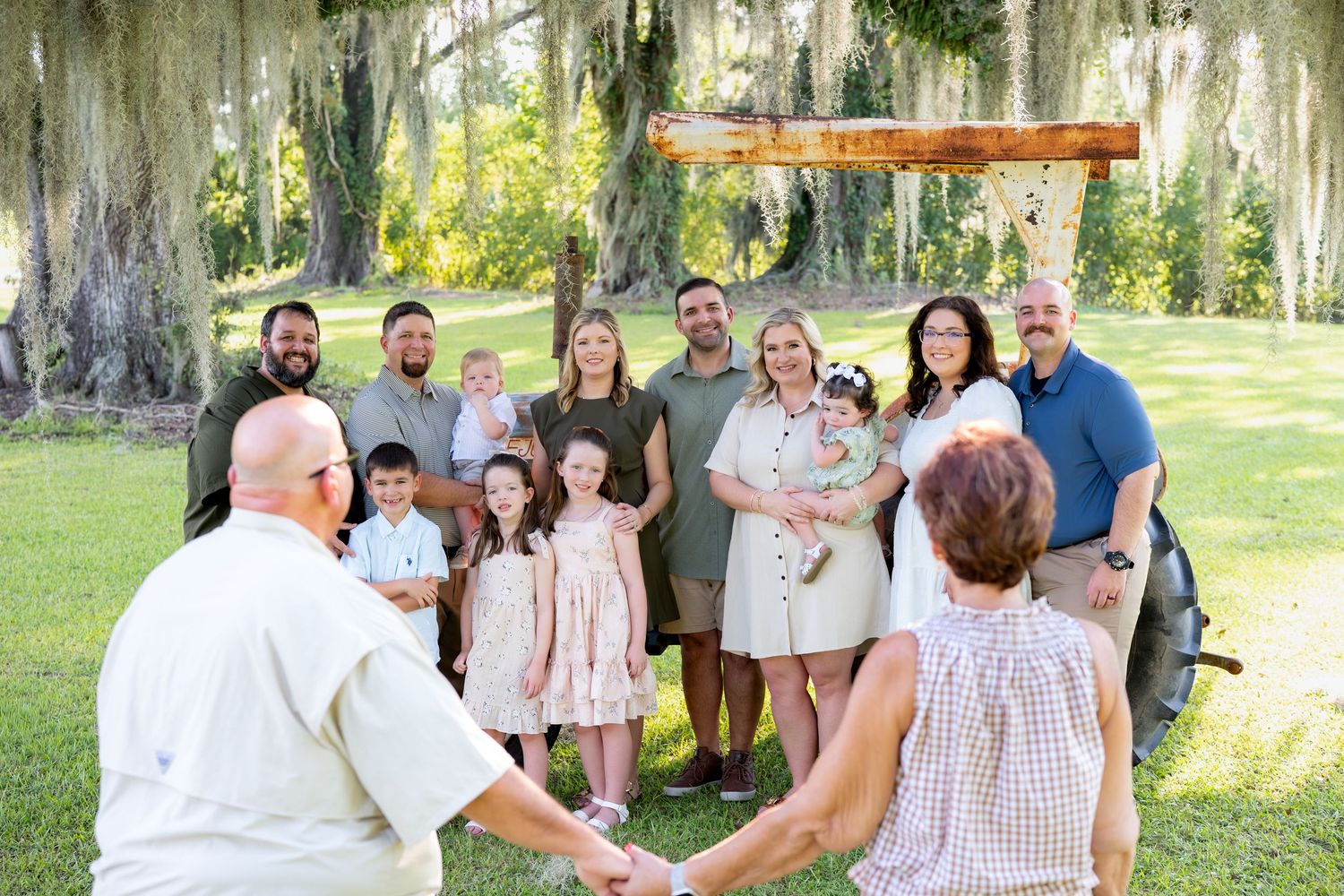 Large family gathering outdoors under Spanish moss trees, posing in a circle on green grass for a portrait.