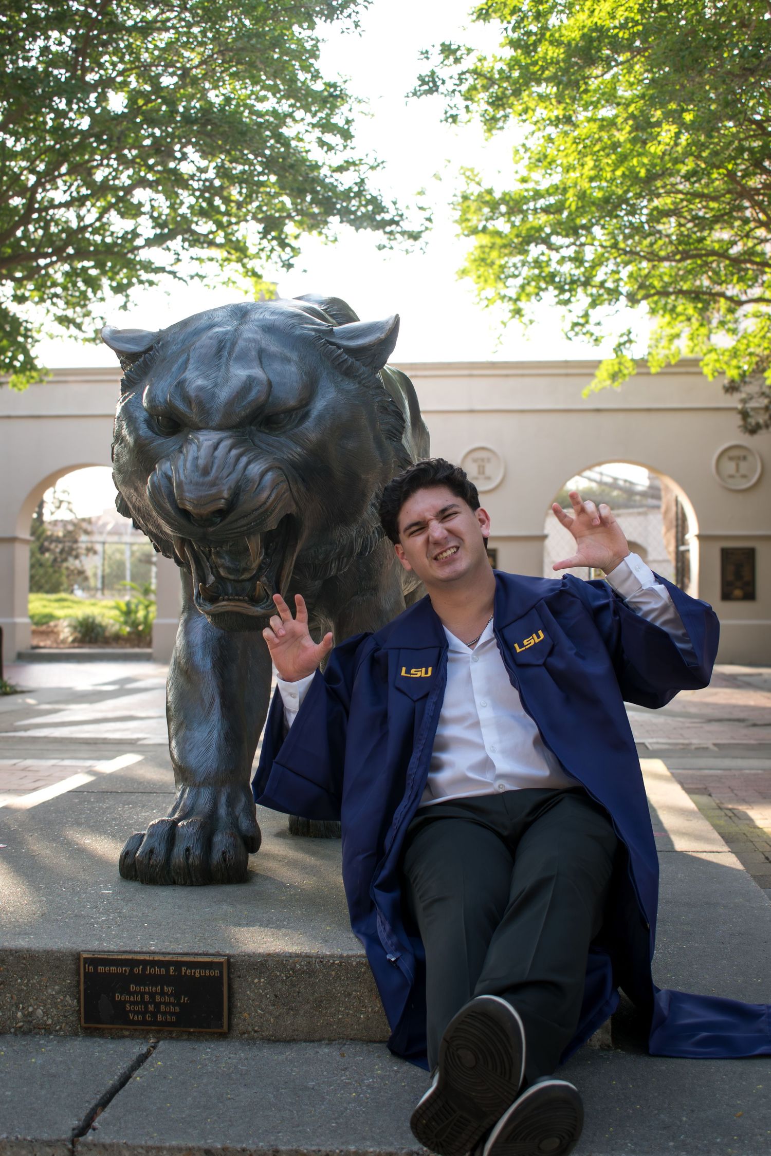LSU graduate in blue gown poses playfully beside bronze tiger statue on campus.