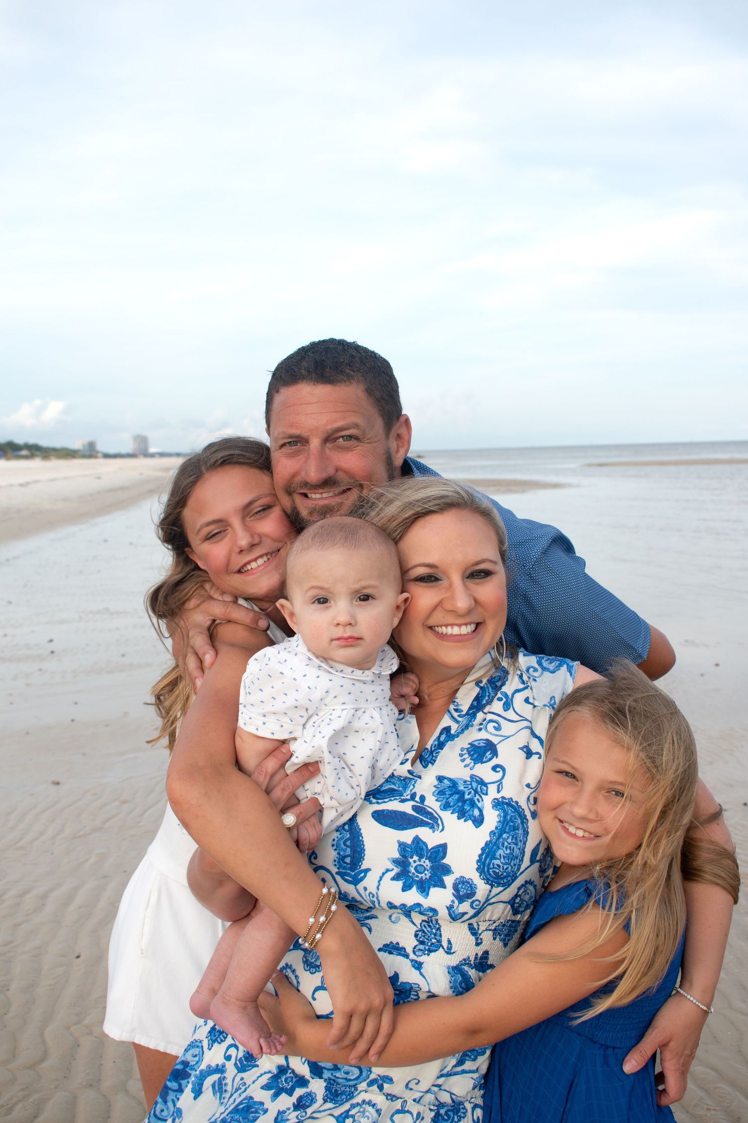 Happy family of five posing together on a sandy beach, with parents holding a baby and two daughters smiling.