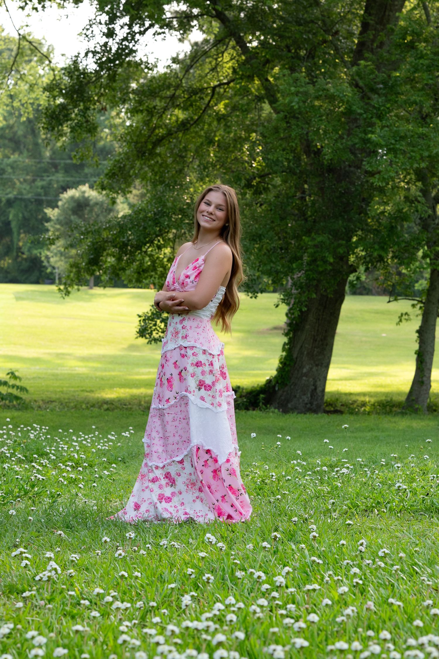 Young woman in pink floral maxi dress standing in green park with white wildflowers and trees.