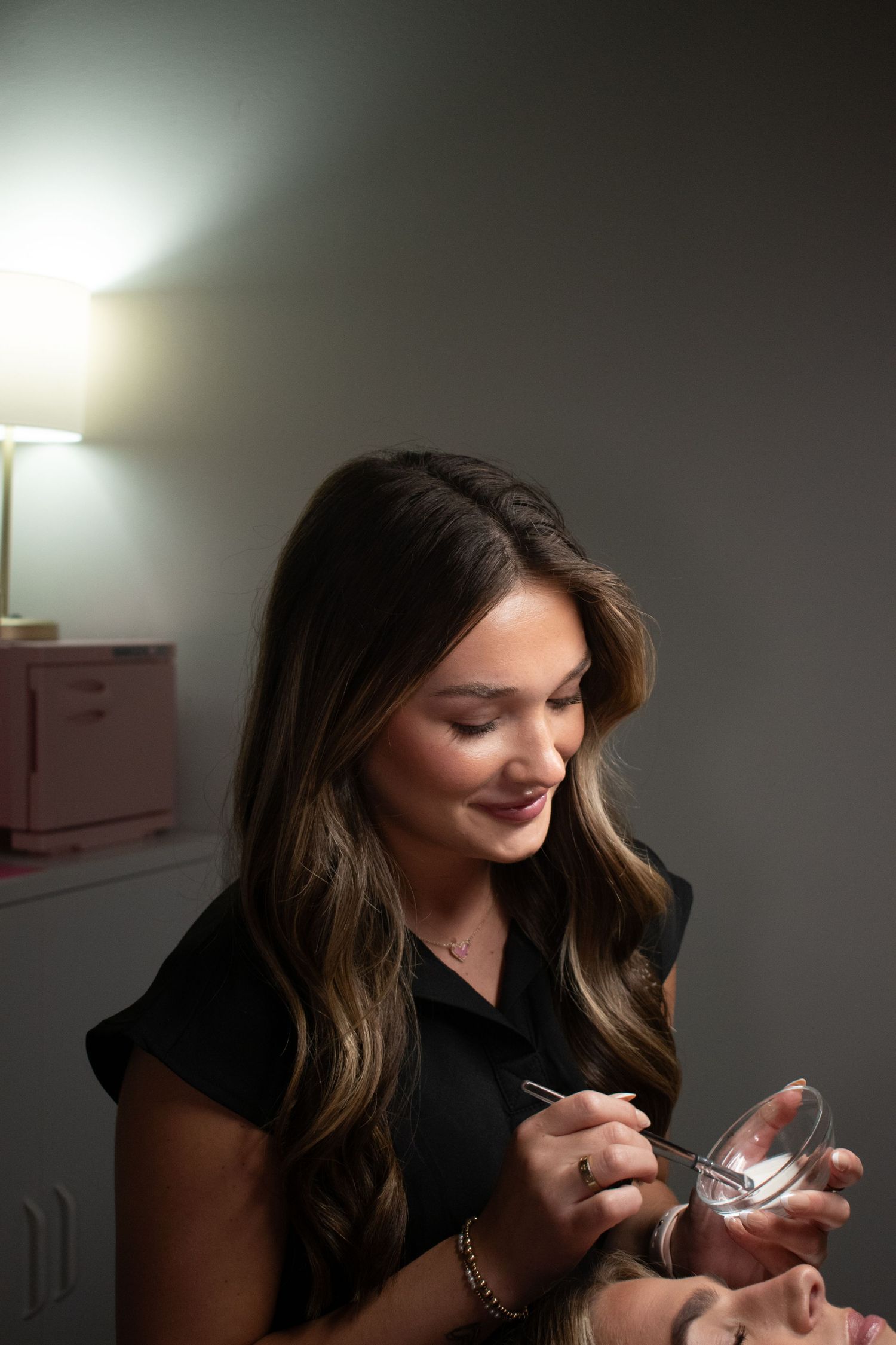 A brunette esthetician in black scrubs carefully applies lash extensions to a client in a dimly lit room.