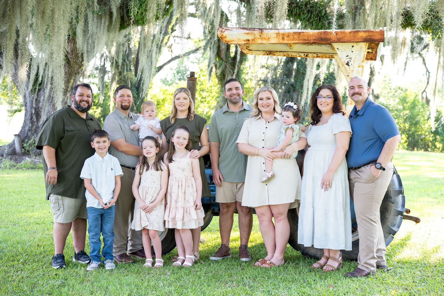 Large family of adults and children posing outdoors on green grass beneath oak trees draped with Spanish moss, beside a vintage tractor.
