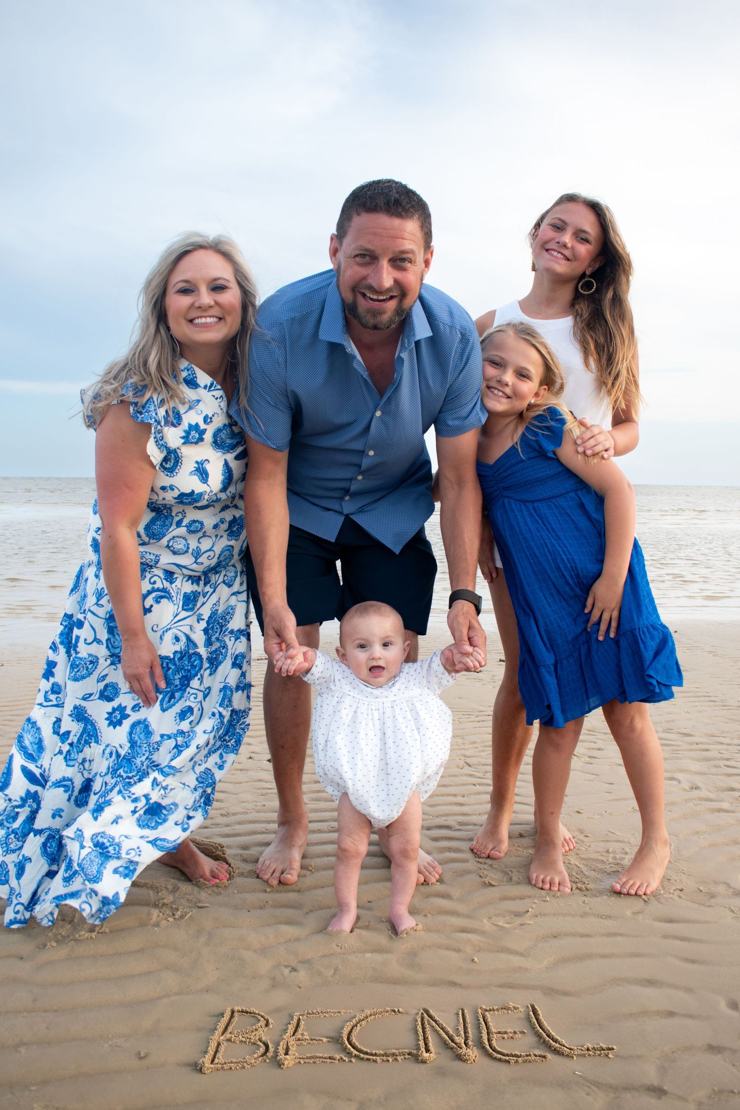 Family of five posing on beach with name BECNEL written in sand, parents with three children including a baby.