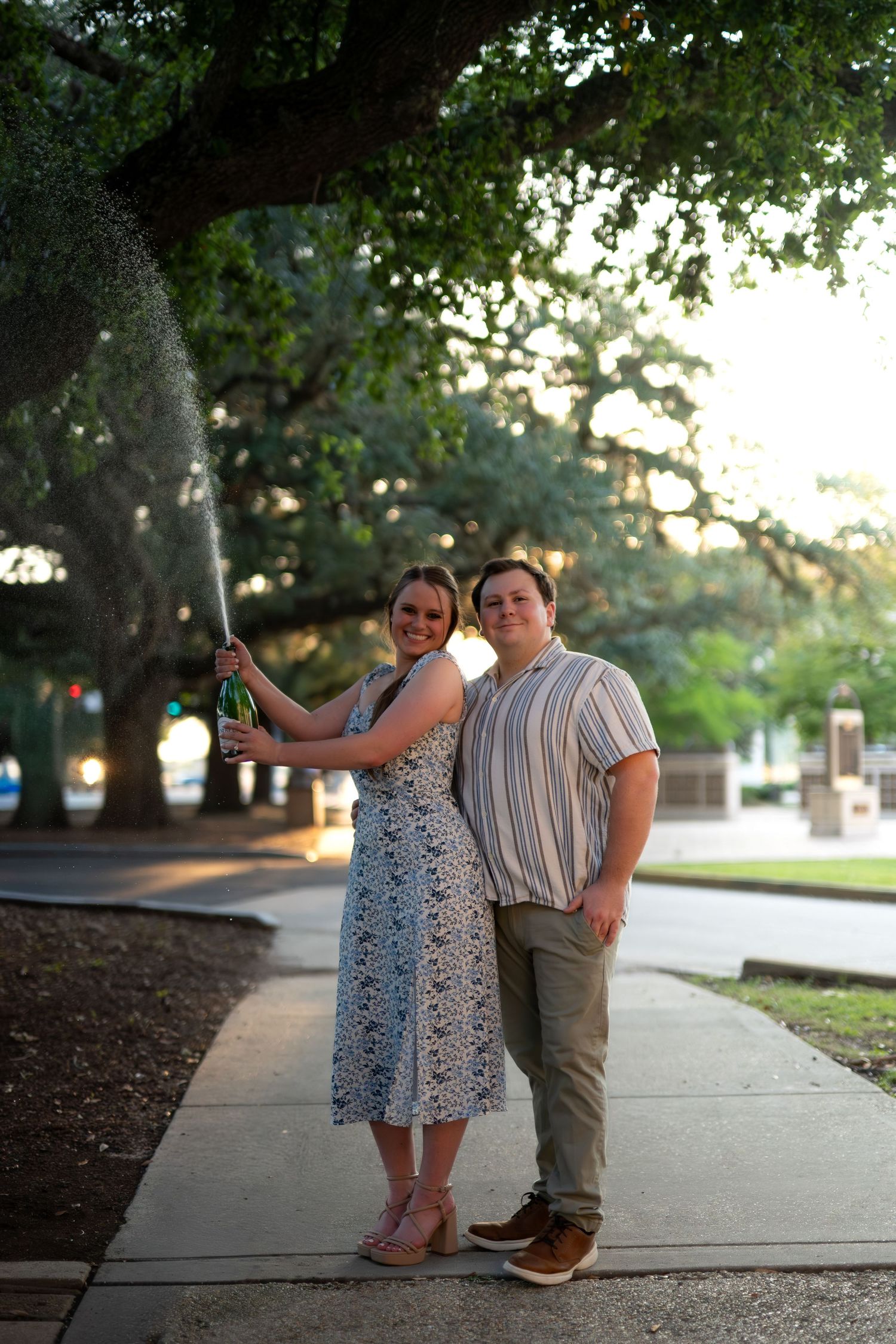 Couple celebrating with champagne spray under large oak trees on a sunny sidewalk.
