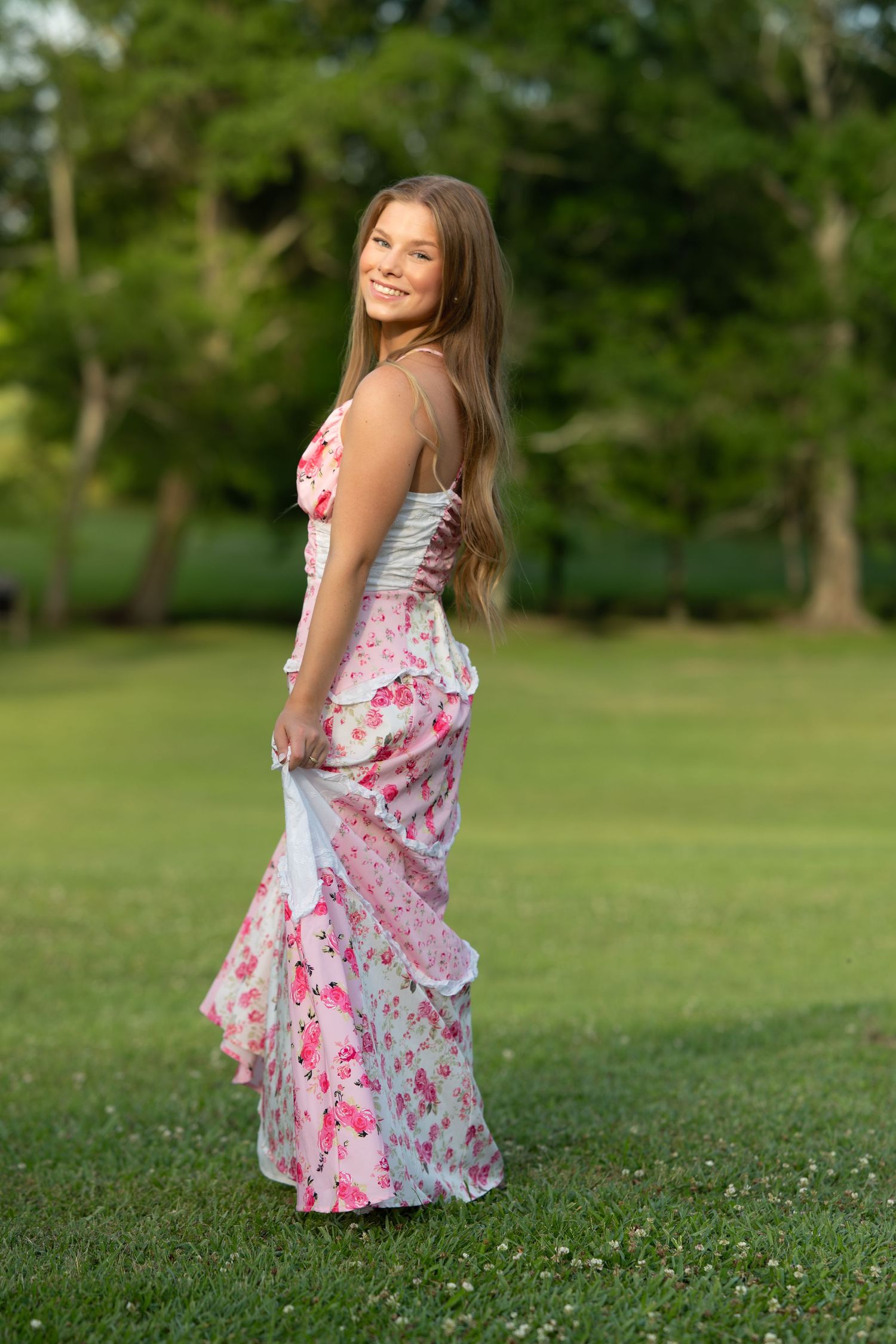 Young woman in pink floral tiered maxi dress twirling on green grass in a sunny park.