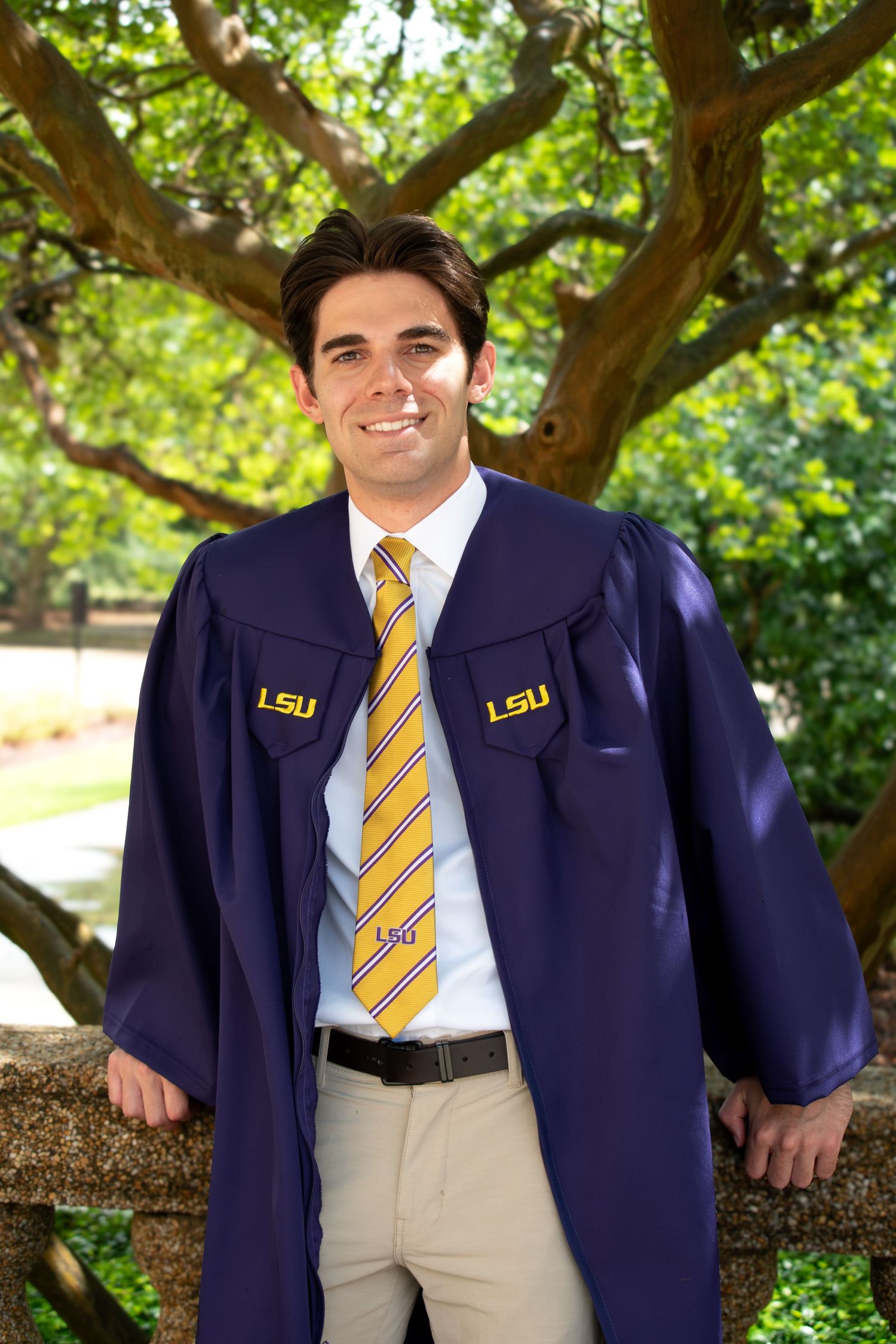Smiling male graduate in LSU navy blue cap and gown with striped tie outdoors.