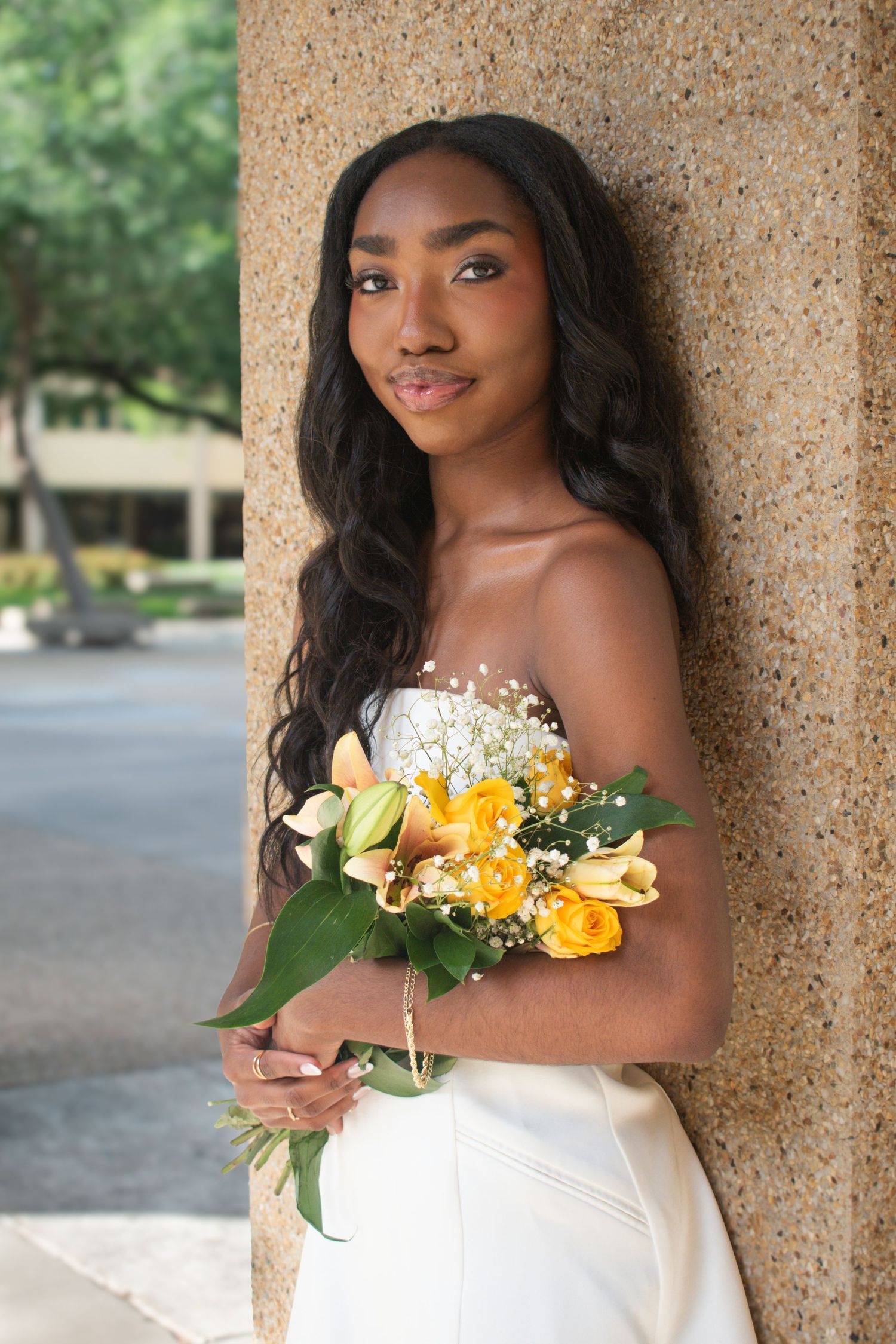 Young woman in white strapless dress holds yellow floral bouquet leaning against stone column.