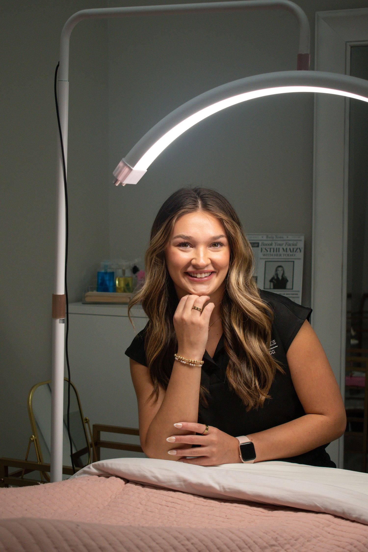 Smiling young woman in black outfit posing under an arc beauty light in a professional waxing studio.