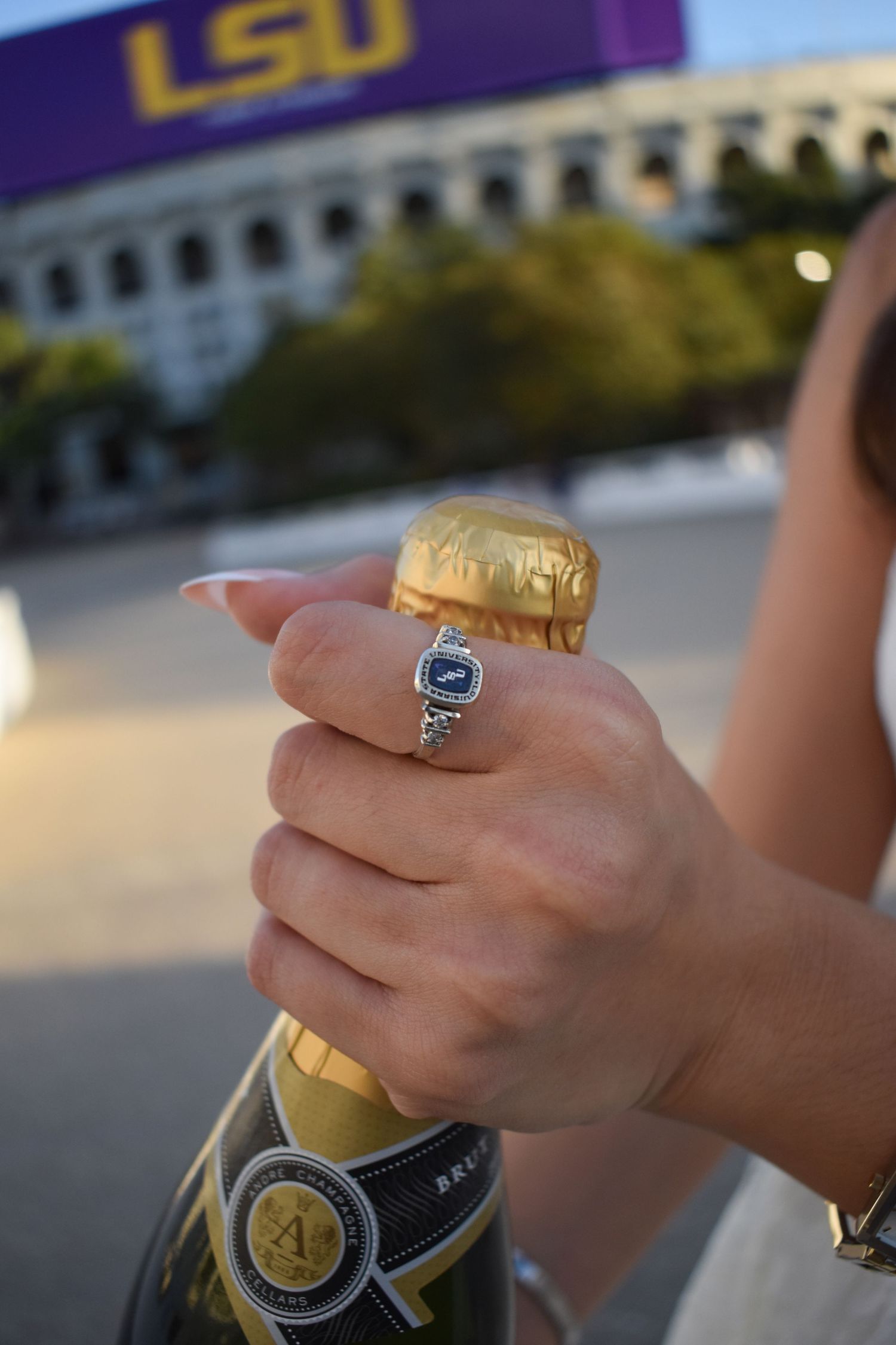 Close-up of hand with sapphire ring opening champagne bottle near LSU stadium at sunset.